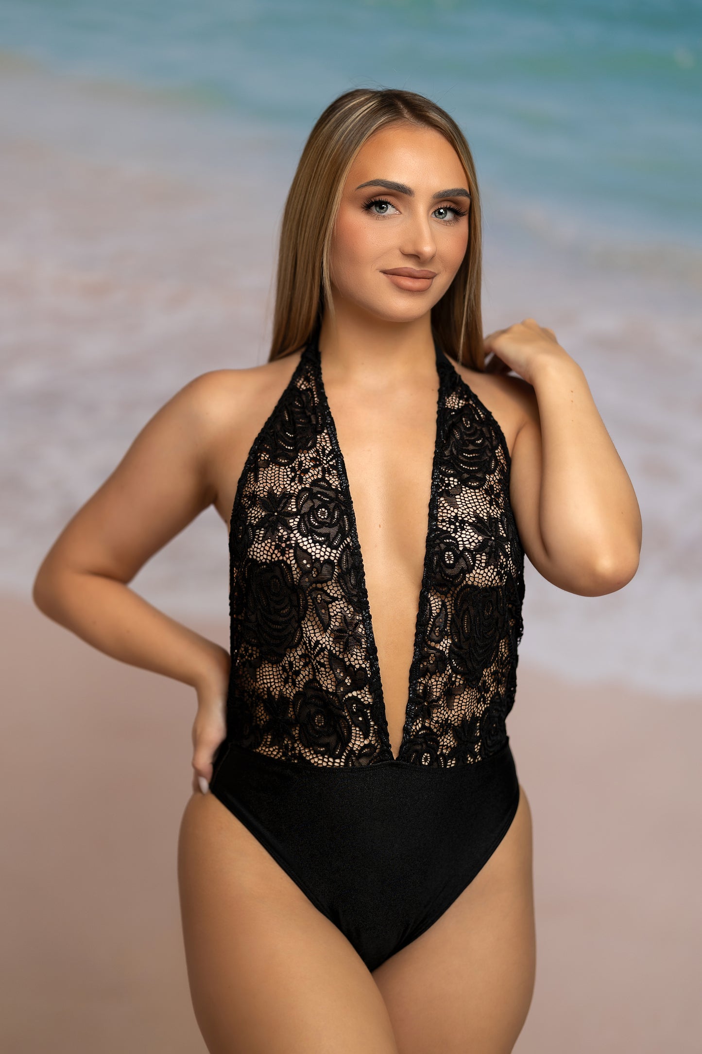 A woman wearing the Brigitewear International High Waist Halter One Piece swimsuit stands on the beach, with waves and sand in the background.