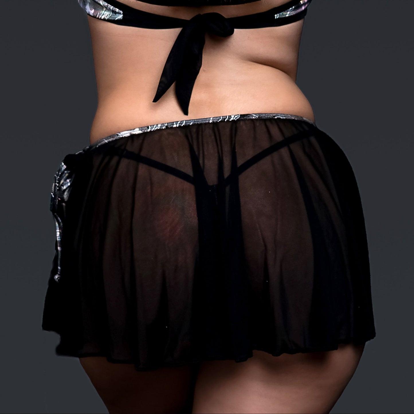 Model in sheer black Electric Glint rave swimwear by Brigitewear International, arms raised and seen from behind against a dark background.