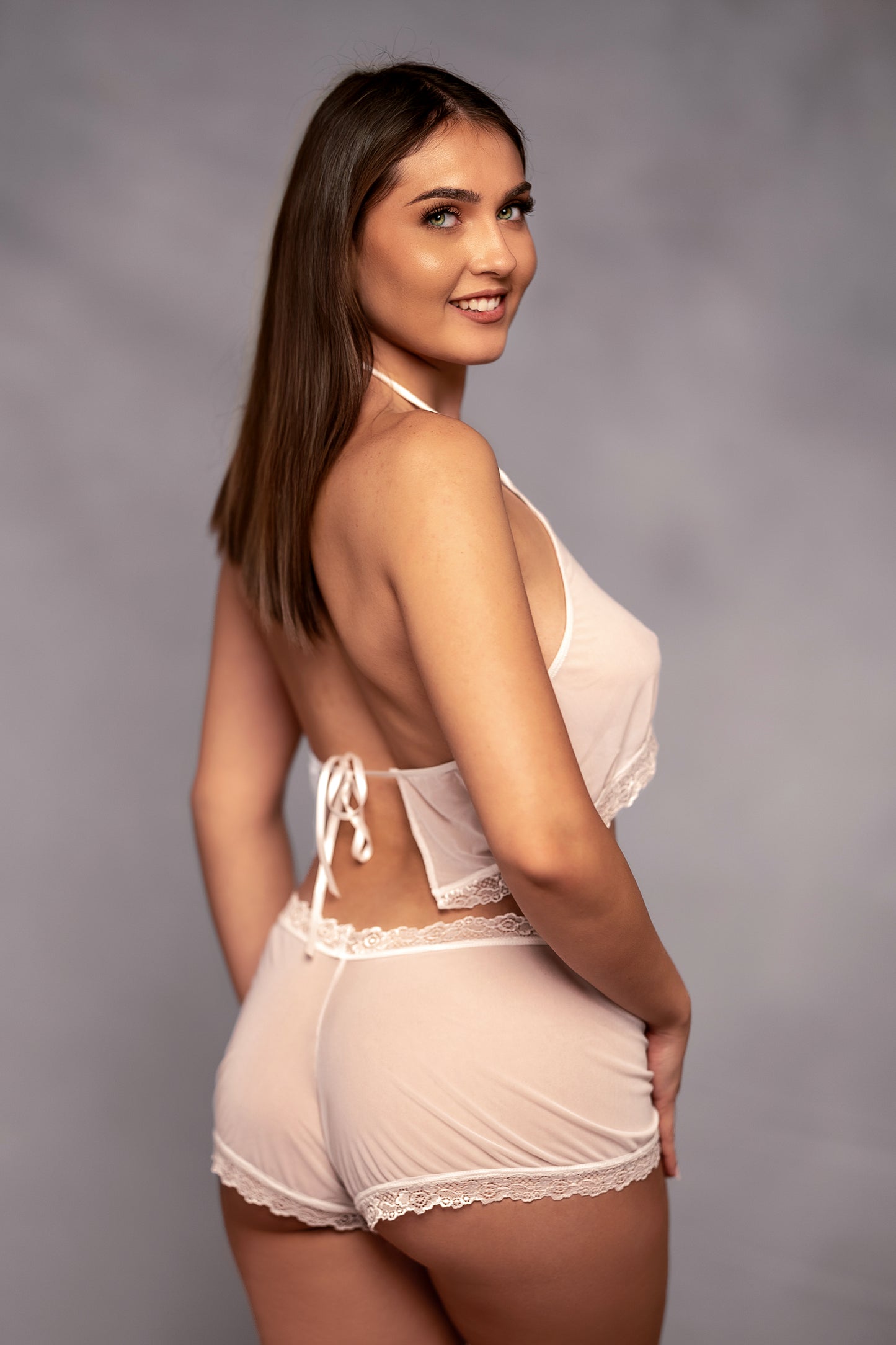 A woman models Brigitewear International's Malibu Nights Boy Shorts in light pink lace, smiling and posing against a gray background while looking over her shoulder.