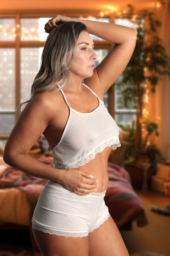 A woman in Brigitewear International's Malibu Nights Boy Shorts stands in a cozy, softly lit bedroom with large windows and lush plants.