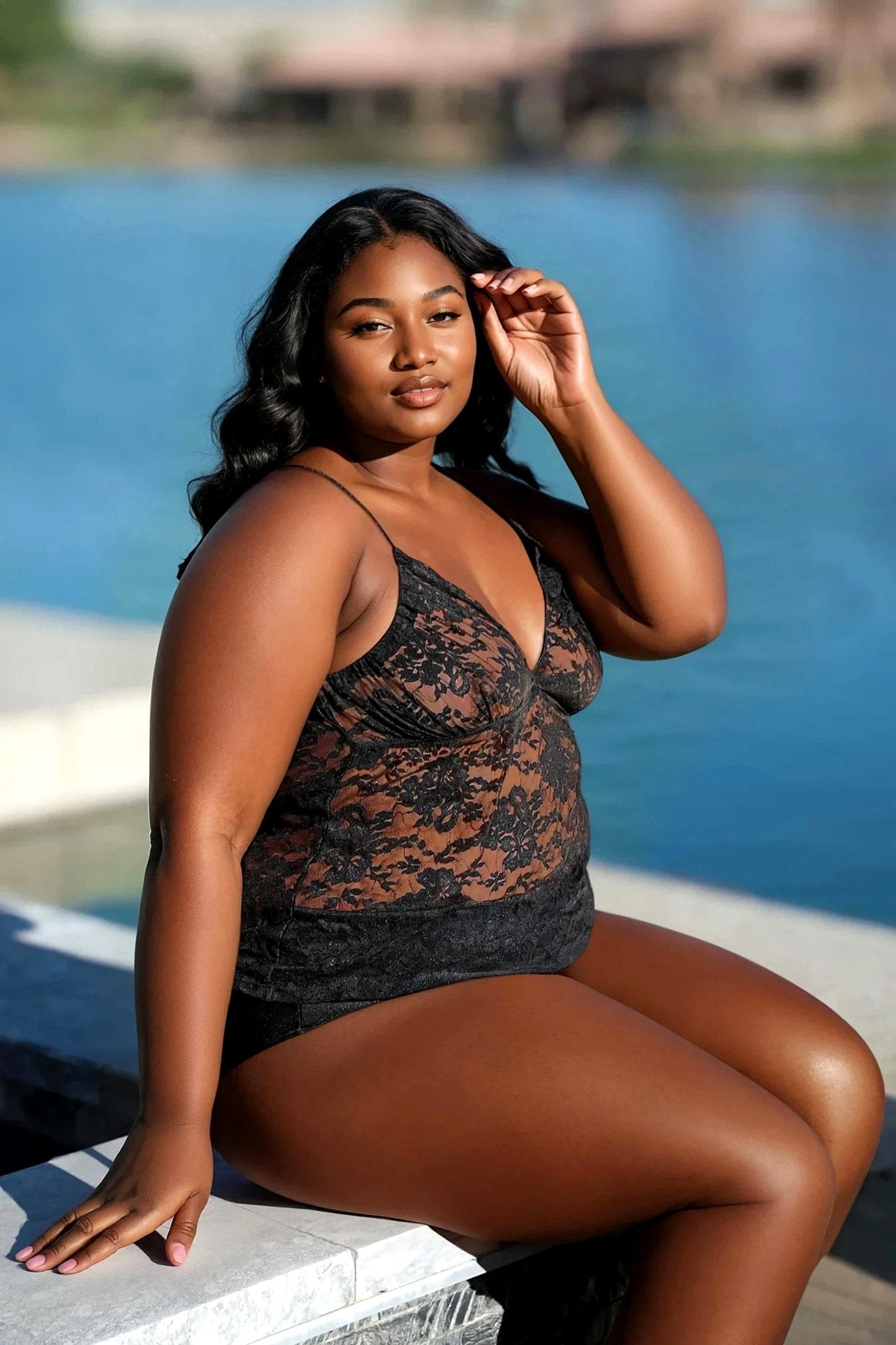 A woman wears the Lovely Lace Halter Sheer Top by Brigitewear International, LLC, sitting by the waterfront and confidently touching her hair while looking at the camera.