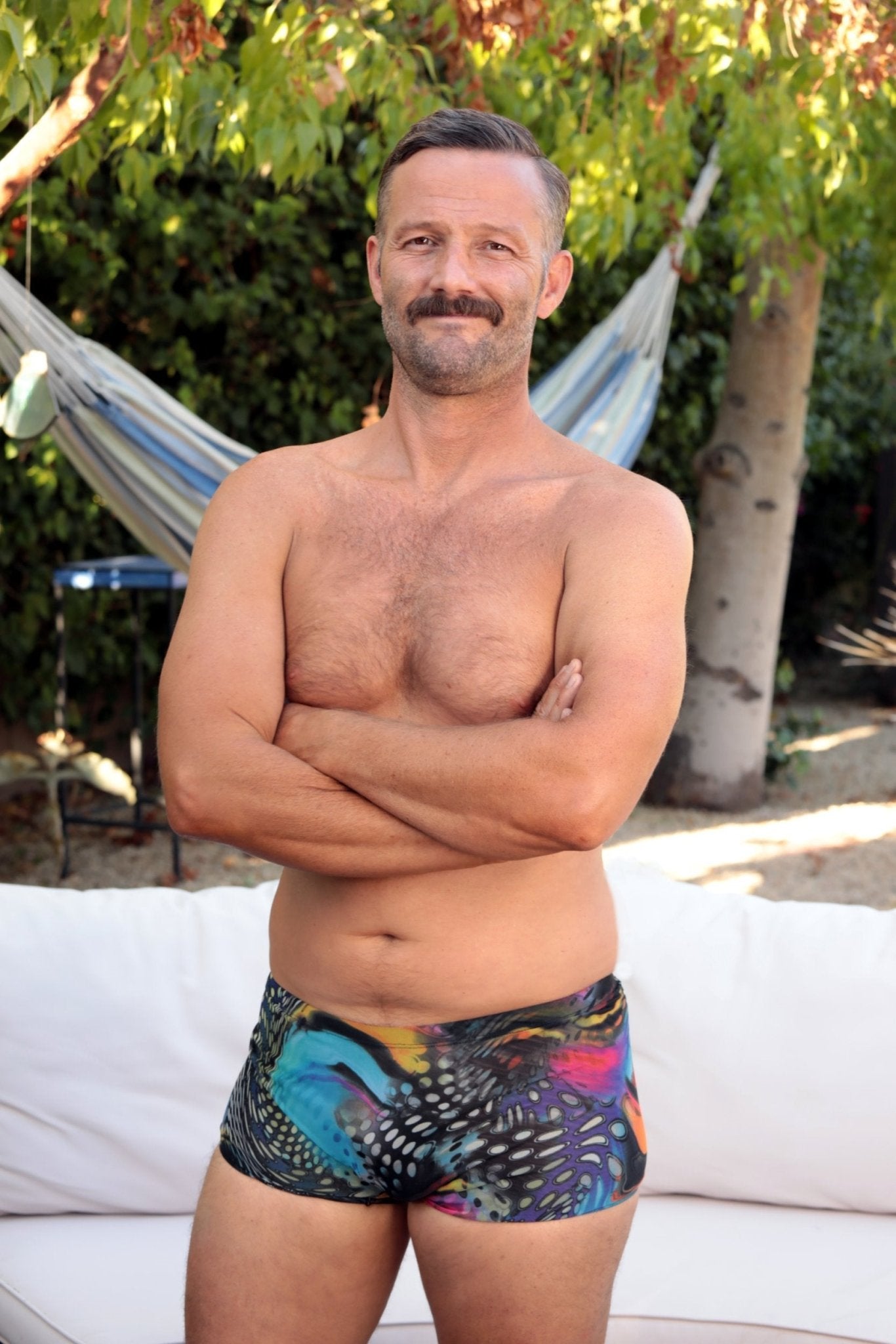A man with a mustache stands outdoors in front of greenery and a hammock, wearing Brigitewear International’s Low Cut Men’s Swimsuit Shorts – Sexy Minimalist Swim Brief.