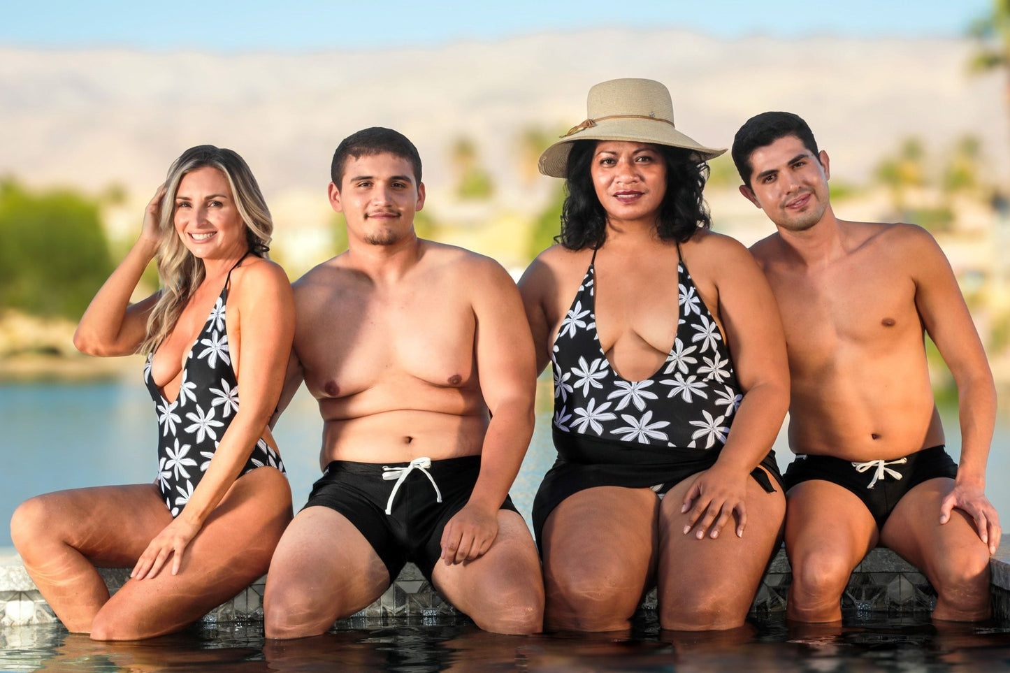 Four people wearing Brigitewear International's Midnight Bloom 1-Piece Swimsuit, featuring a stunning floral design, sit poolside and smile at the camera.