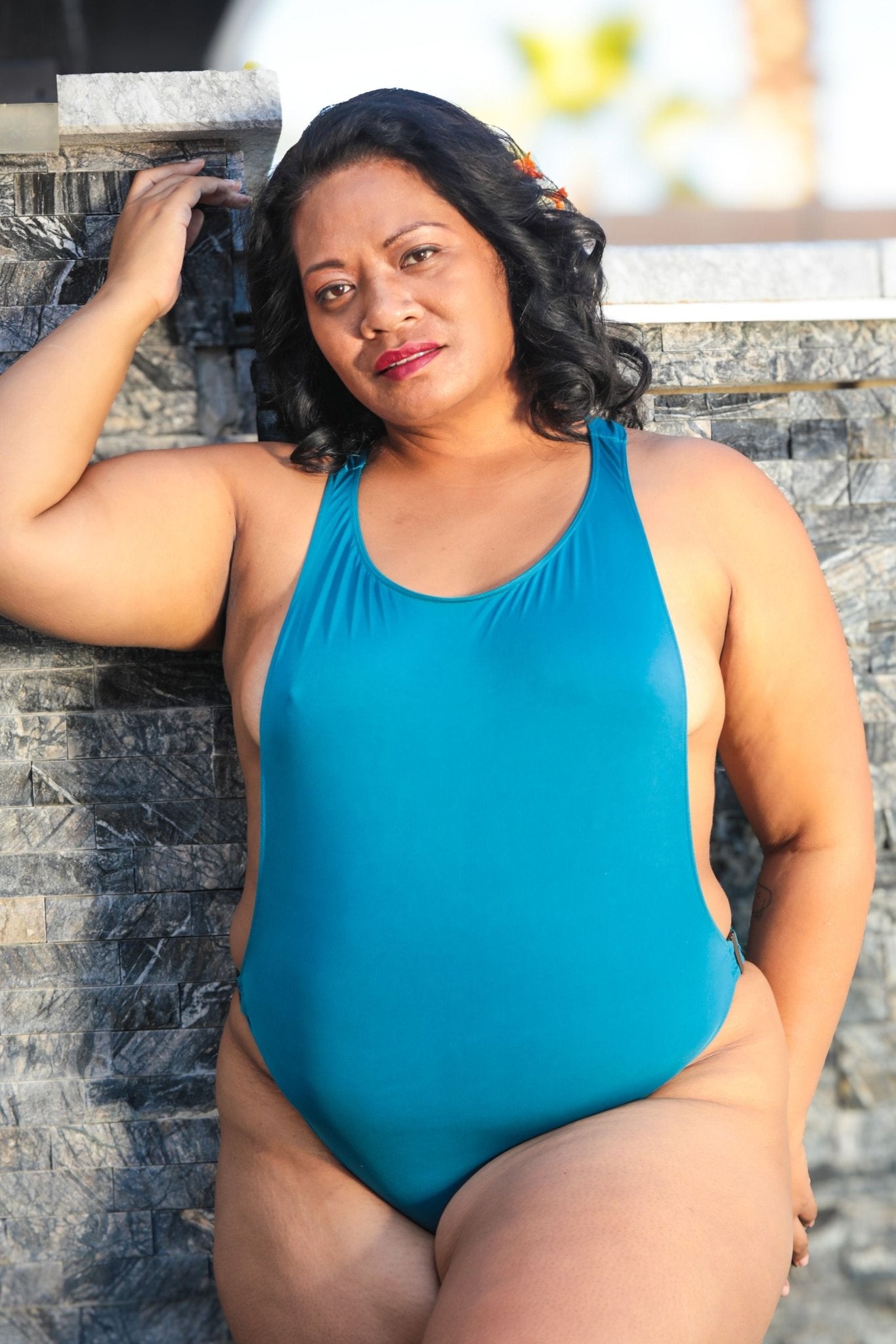 A woman models the Peek-a-Boob One-Piece Swimsuit by Brigitewear International, LLC, leaning against a stone wall and gazing at the camera with a relaxed expression.