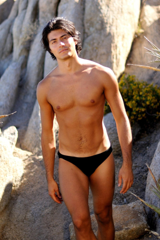 A man wears the Brigitewear International, LLC Speedster Men’s Swimsuit – Classic European Style Swim Brief, standing outdoors among rocks and plants on a sunny day.
