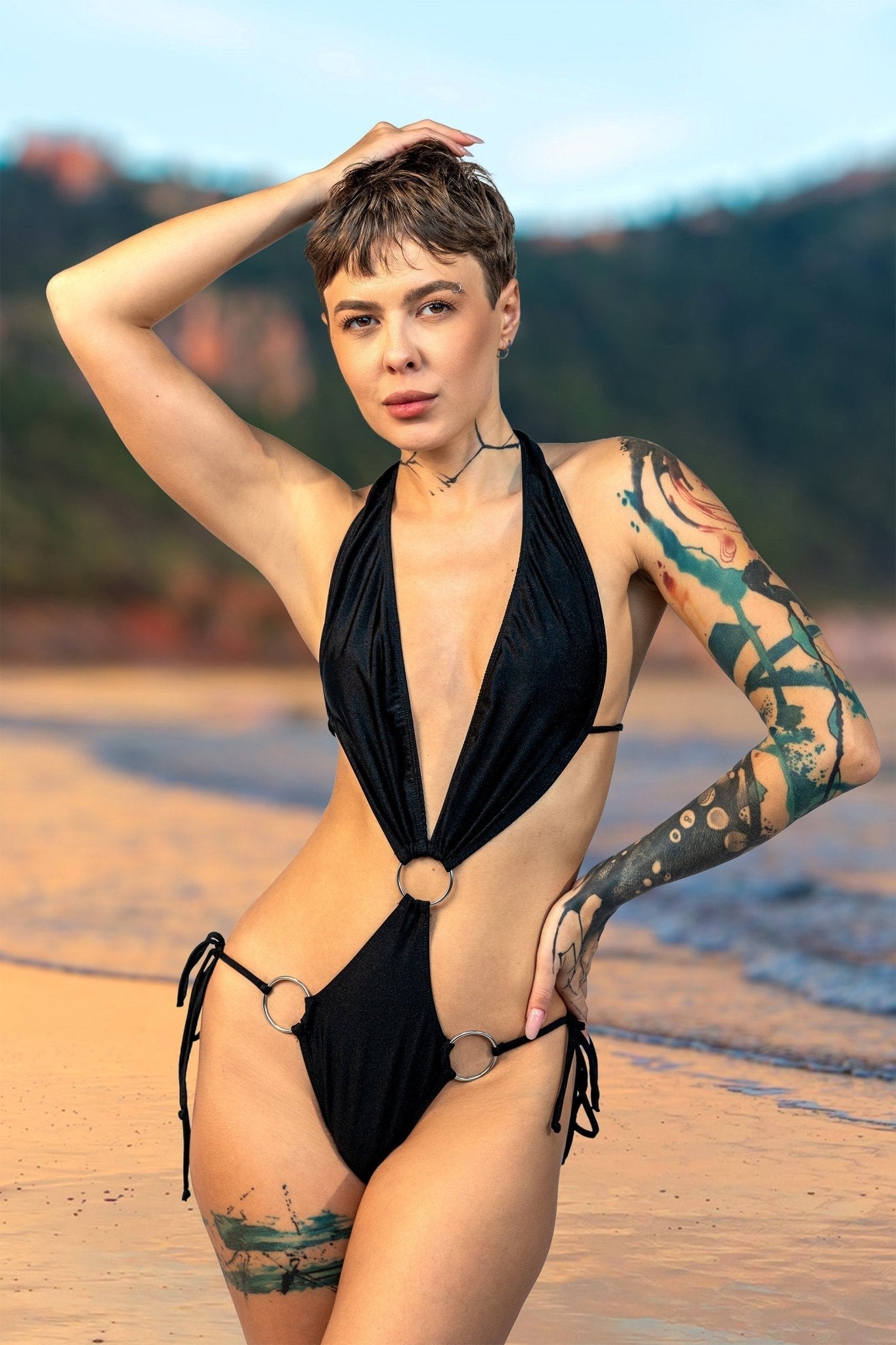 Person with short hair and tattoos posing on a beach in a black swimsuit, with hills in the background.