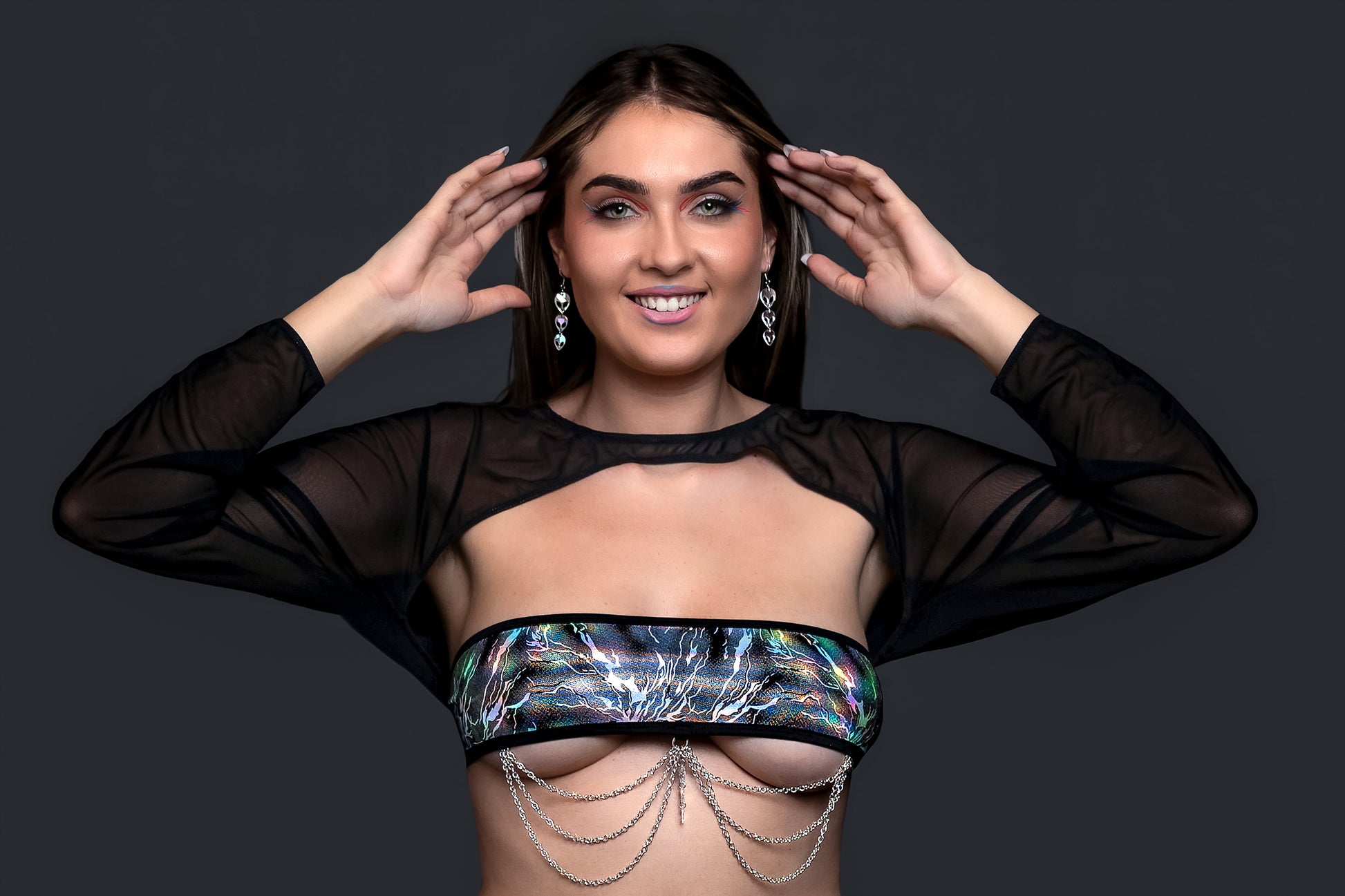 A woman poses with hands near her head, wearing Brigitewear International’s Electric Glint sheer black top and metallic chain bandeau—perfect rave swimwear and festival bikini style.