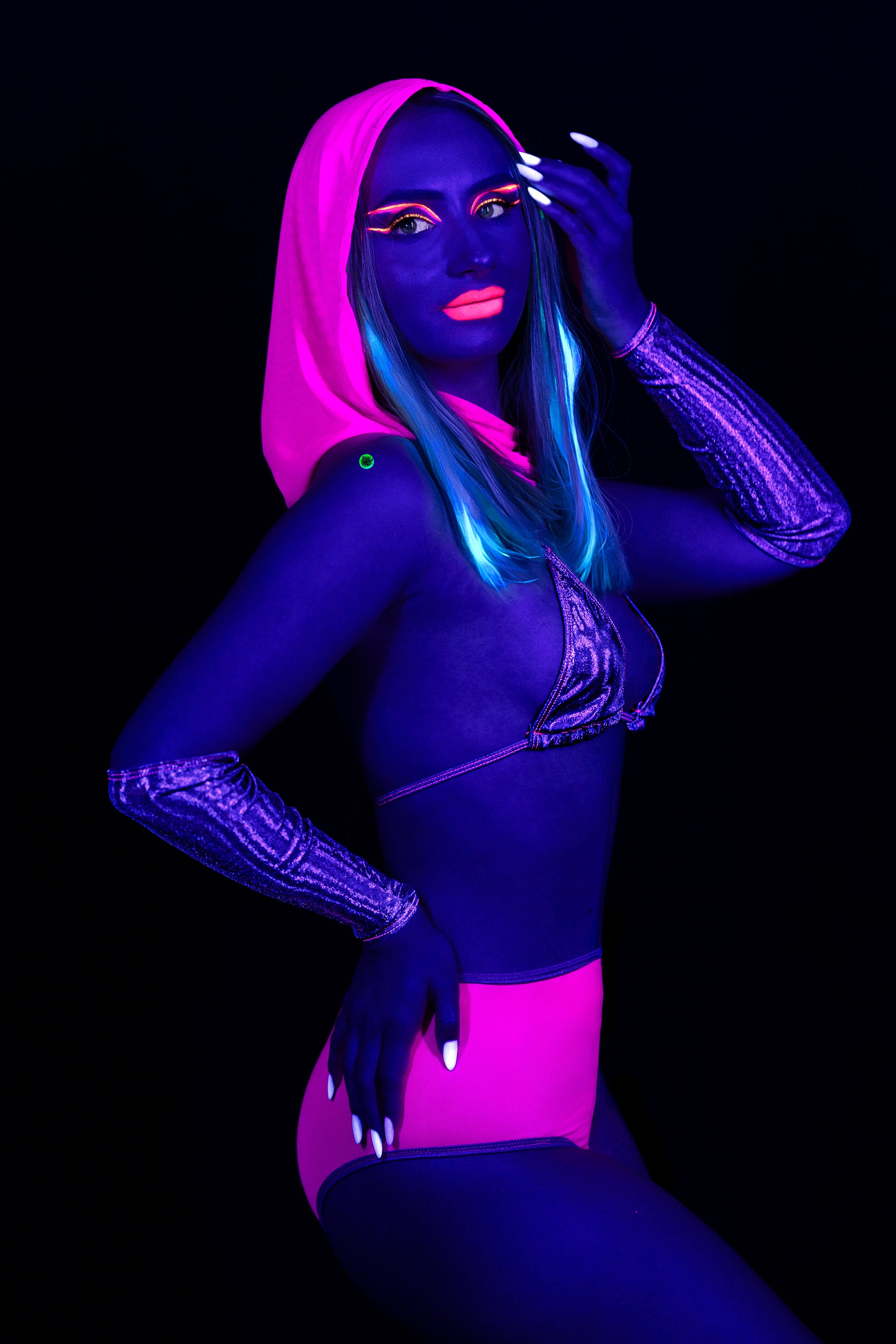 A woman wears the Brigitewear International, LLC Grape Babe Mesh Hood and a neon pink bikini with glowing body paint and hair under blacklight on a dark background.