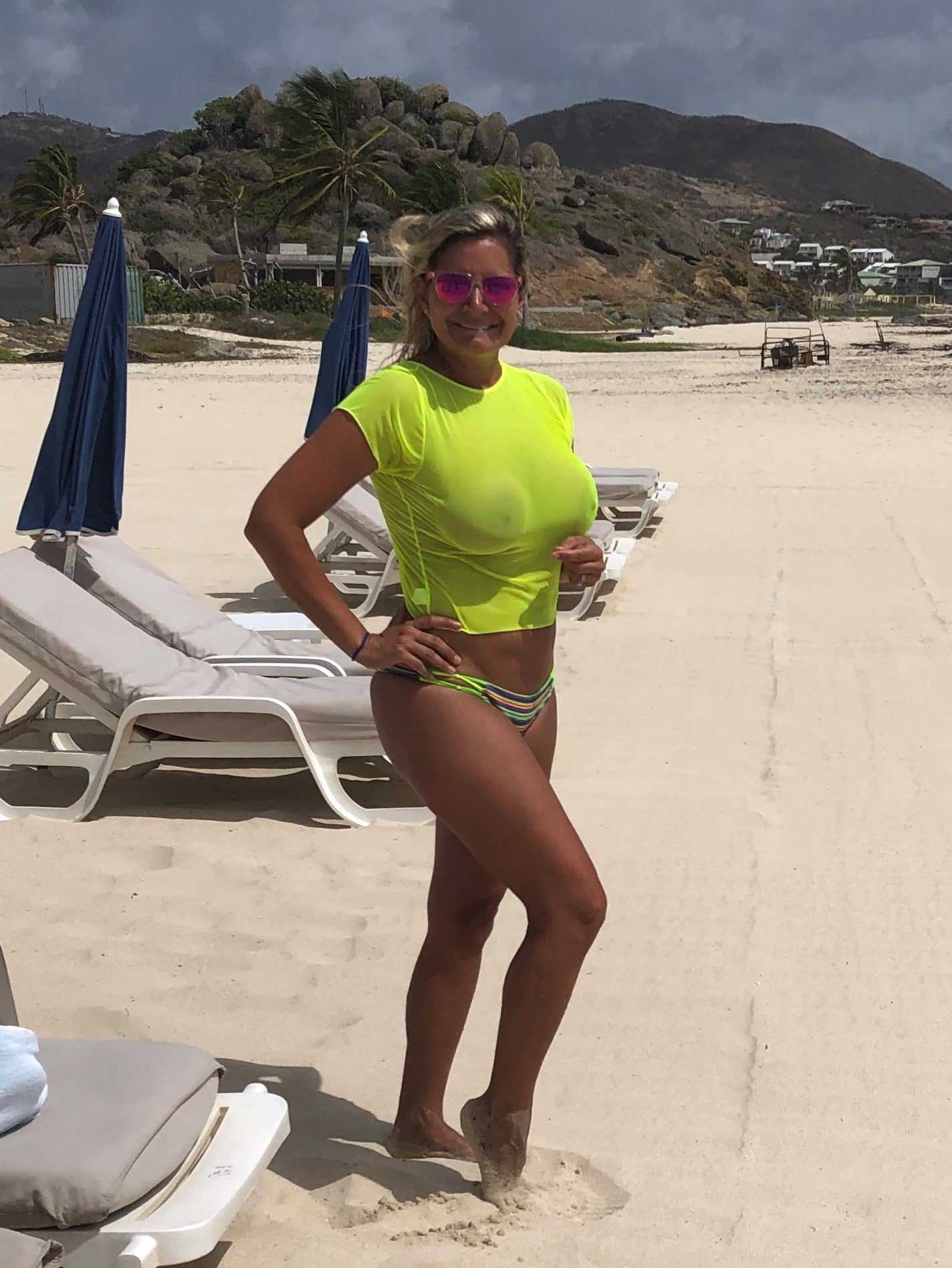 Woman in a neon yellow top and bikini bottoms standing on a sandy beach near lounge chairs and umbrellas.