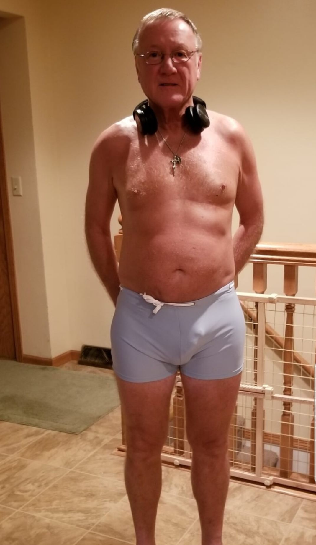 Older man wearing light swim trunks and headphones around neck stands indoors by a staircase and baby gate.