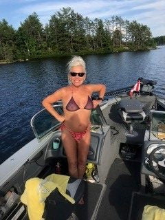 Woman in sunglasses and bikini standing on a boat, smiling, with trees and water in the background.
