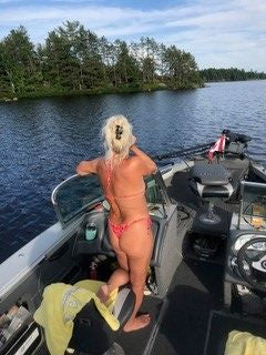 A woman in a red Brigitewear swimsuit stands on a boat, looking out over a calm lake surrounded by trees.