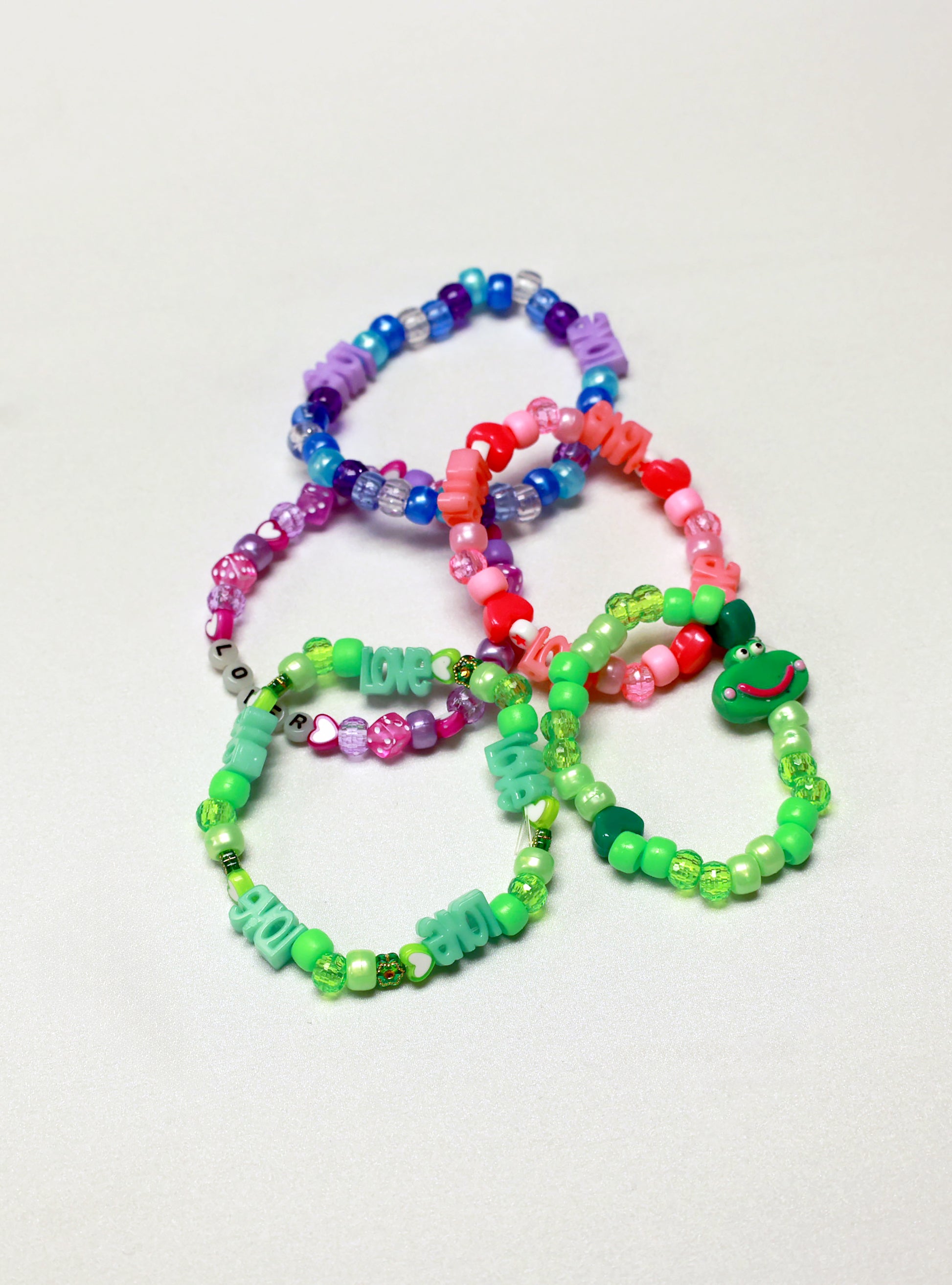 Four vibrant Baby's 1st Kandi bracelets by Brigitewear International, LLC, each featuring letter beads spelling LOVE, are arranged on a white surface.