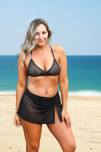 A woman models a black mesh bikini and skirt by Brigitewear International, LLC as she stands on a sandy beach with the blue ocean in the background.