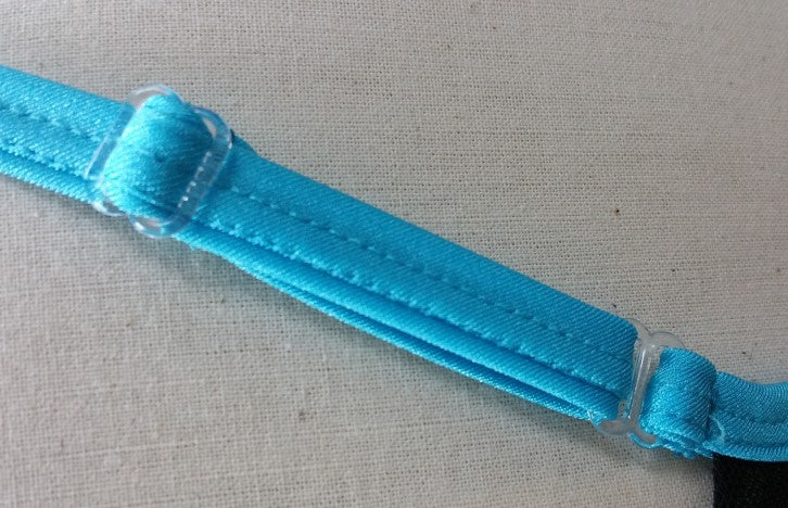 Close-up of a blue adjustable bra strap with plastic sliders, part of a supportive mesh bra by Brigitewear International, LLC, set against a beige fabric background.