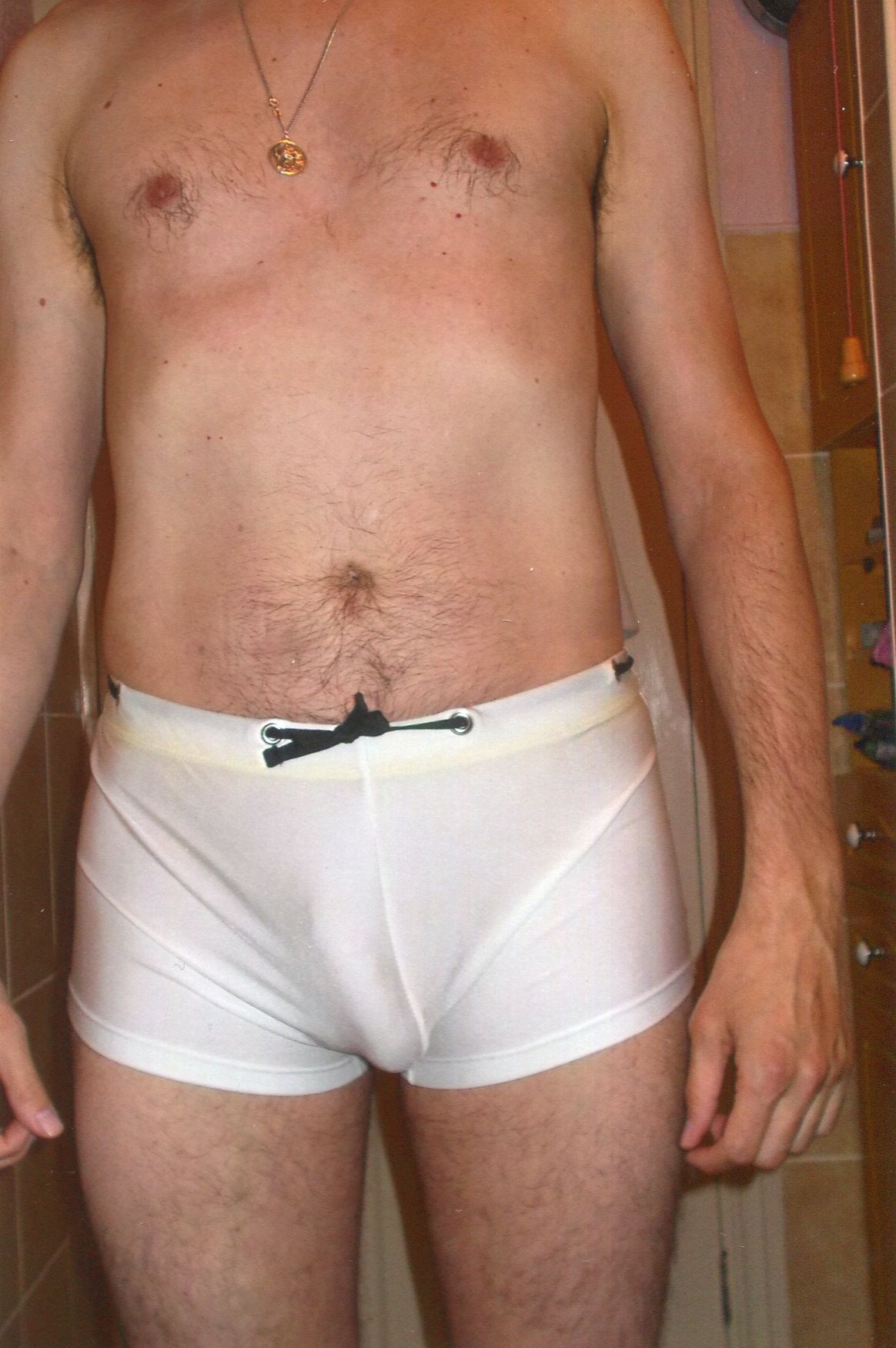 A man wearing white boxer briefs, reminiscent of sexy swimwear, stands indoors with his torso and upper legs visible.