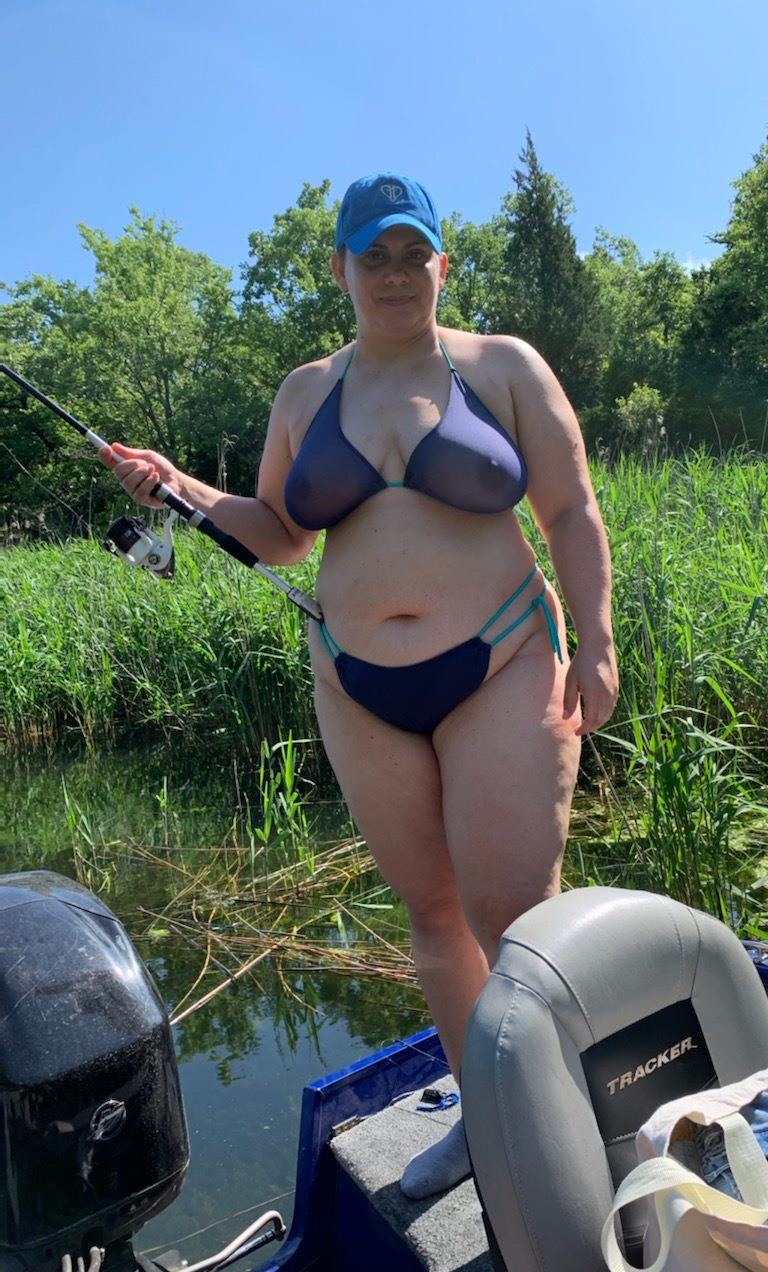Woman in sexy swimwear—a blue bikini and cap—fishing on a boat surrounded by tall grass and trees on a sunny day.