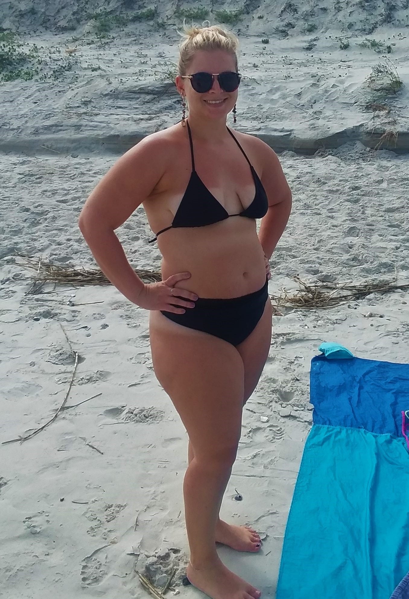 Woman in black Brigitewear bikini and sunglasses stands on a sandy beach with towels on the ground nearby, embodying elegant resort wear style.