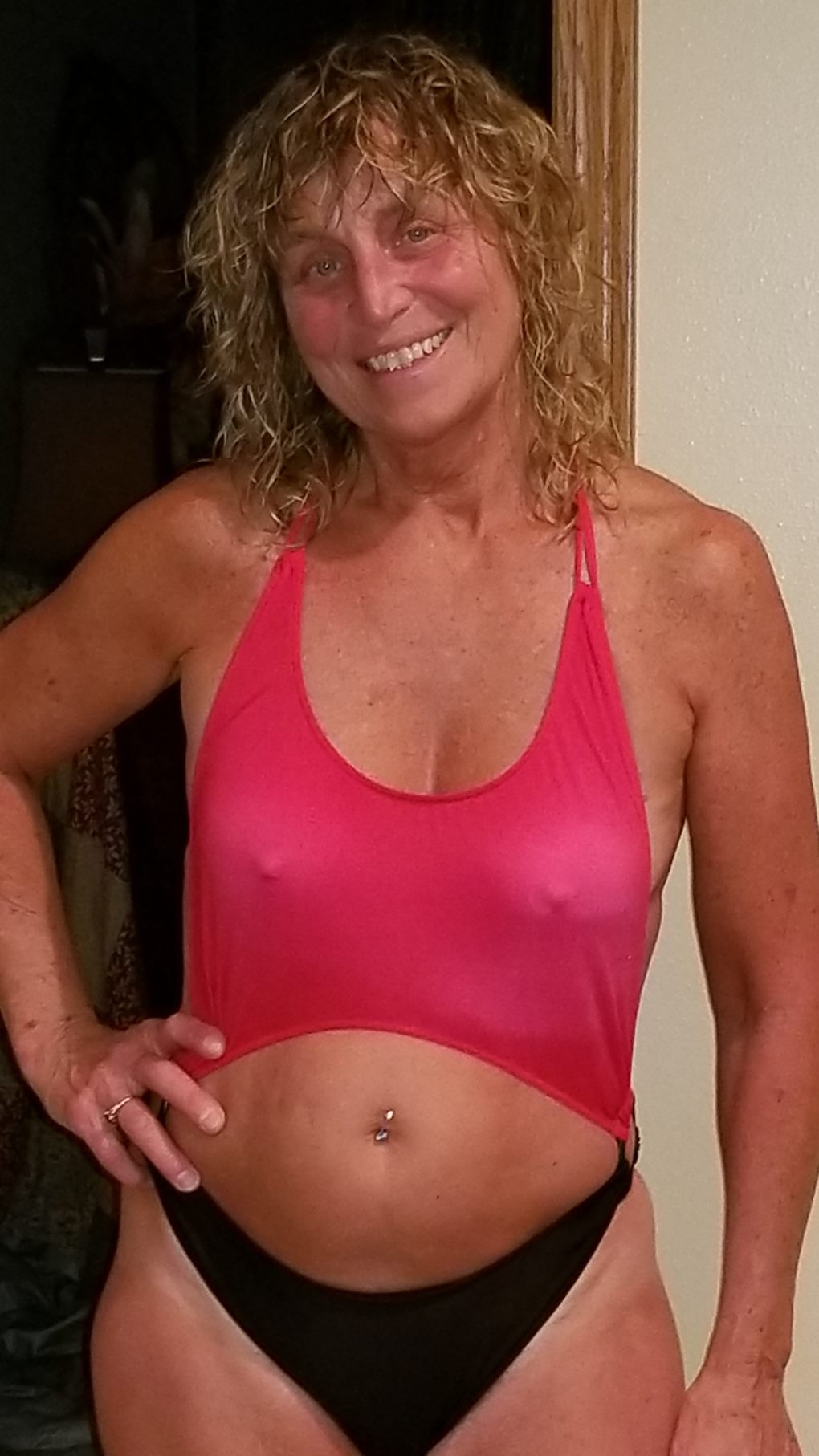 Smiling woman with wavy hair wearing a red and black Brigitewear swimsuit, standing indoors with one hand on her hip.