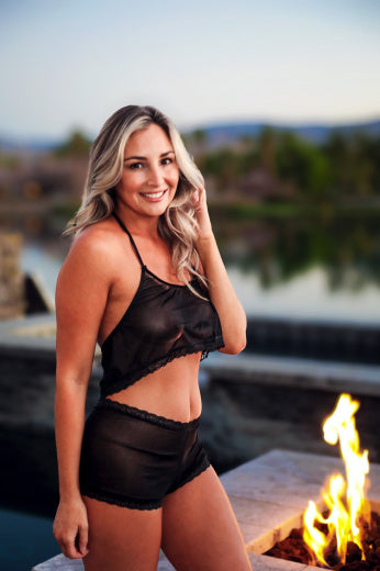 A woman wearing Brigitewear International's Malibu Nights Boy Shorts smiles by a fire pit outdoors, with a lake and trees in the background.