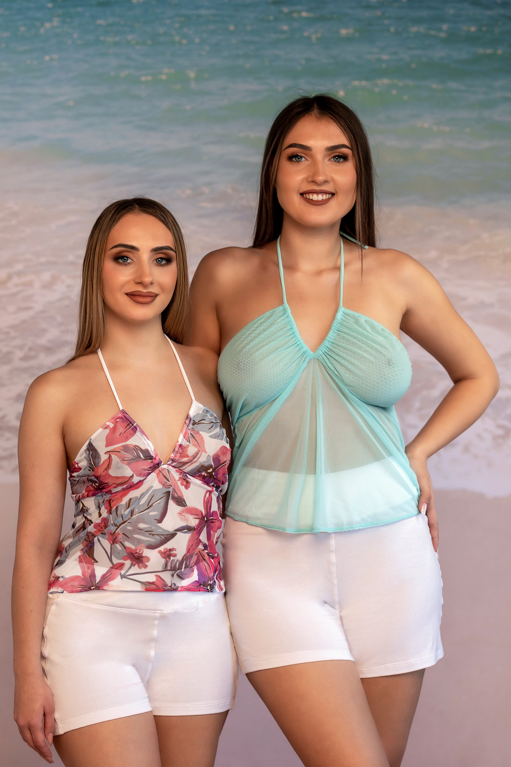 Two women wearing Brigitewear International, LLC’s Plunge Top stand on a beach with ocean waves behind them, smiling at the camera.