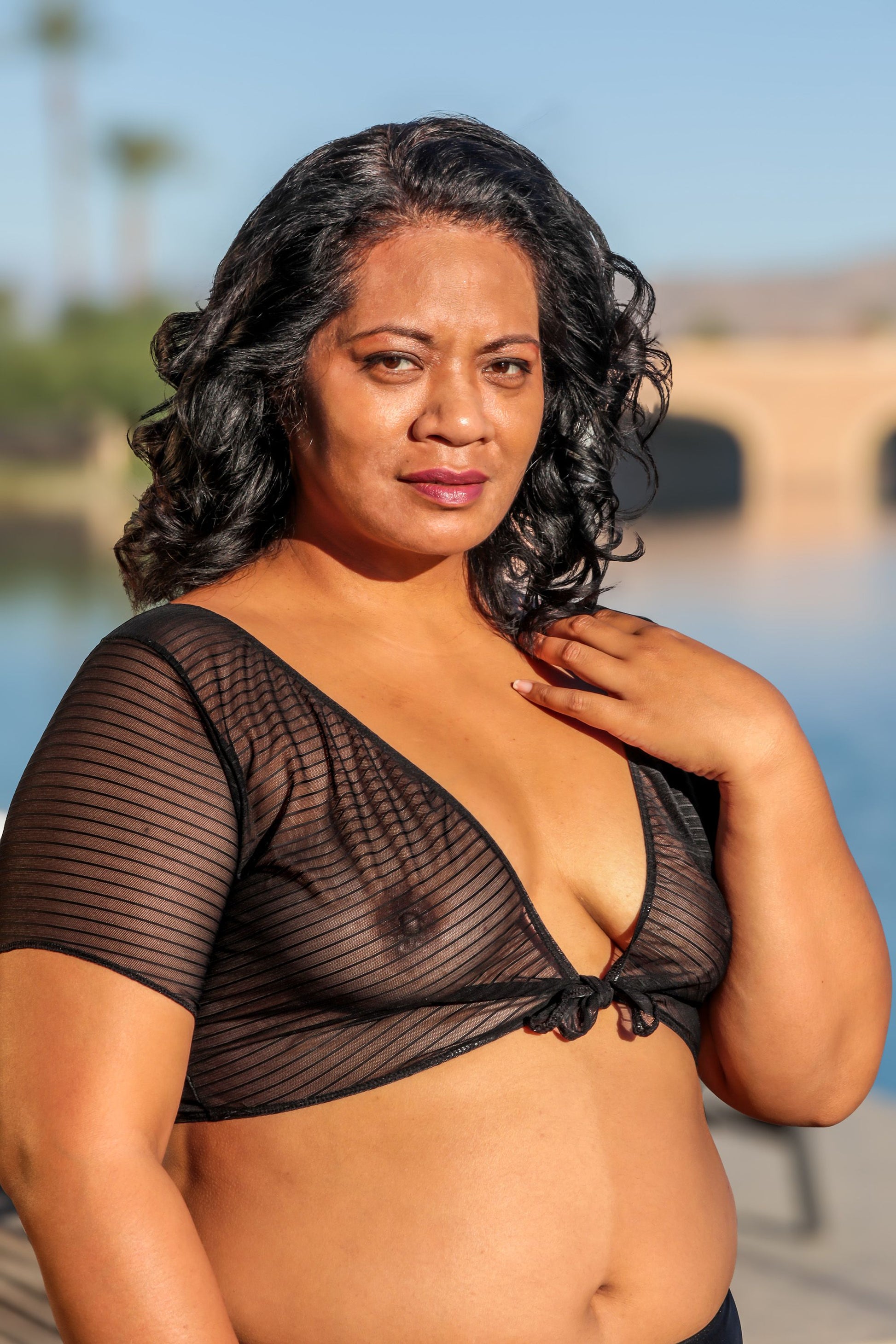 A woman wearing the Brigitewear International, LLC Sheer TieTee stands outdoors by the water, looking at the camera.