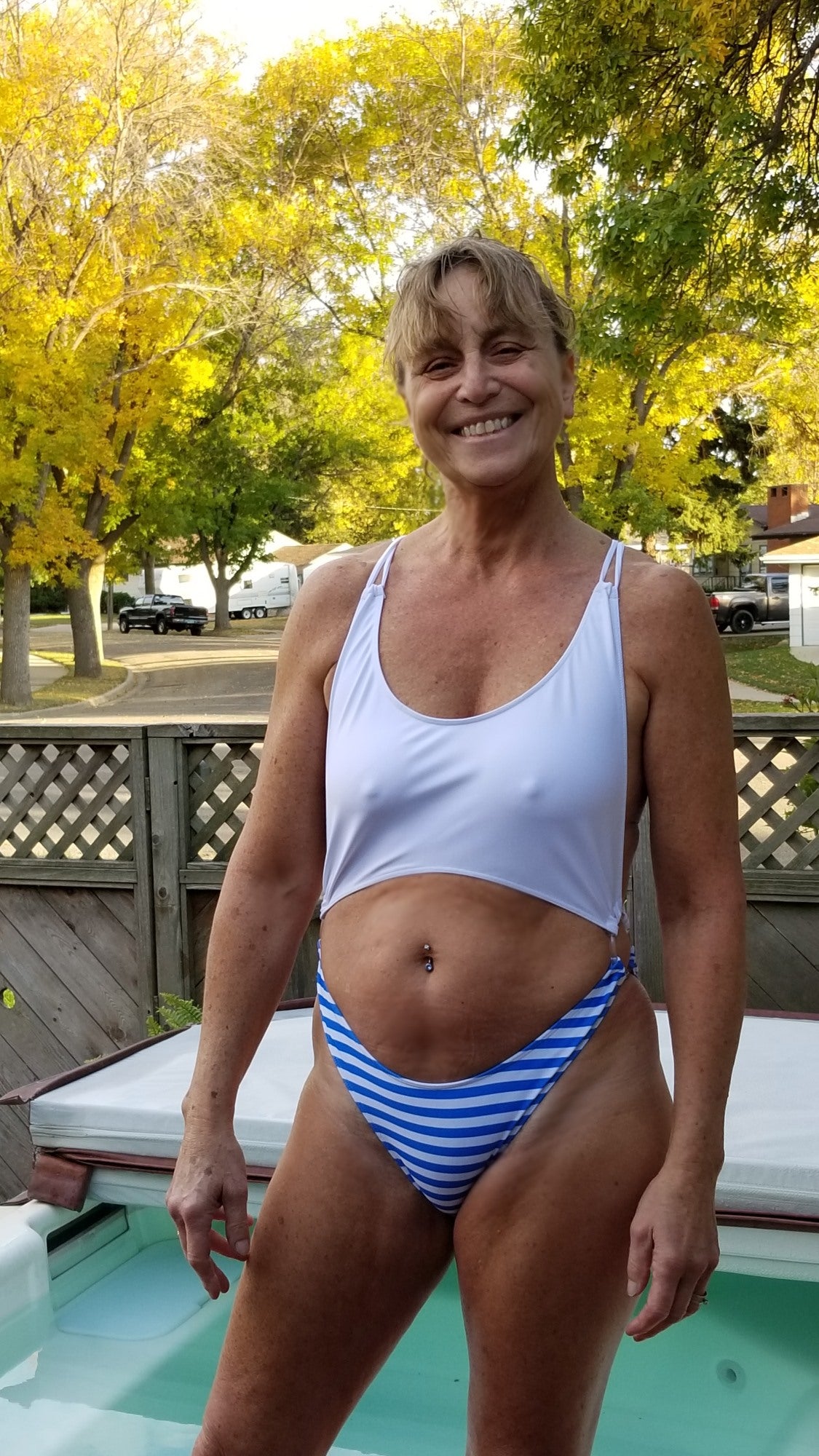Smiling woman in a Brigitewear bikini stands by a pool with trees and a wooden fence in the background.