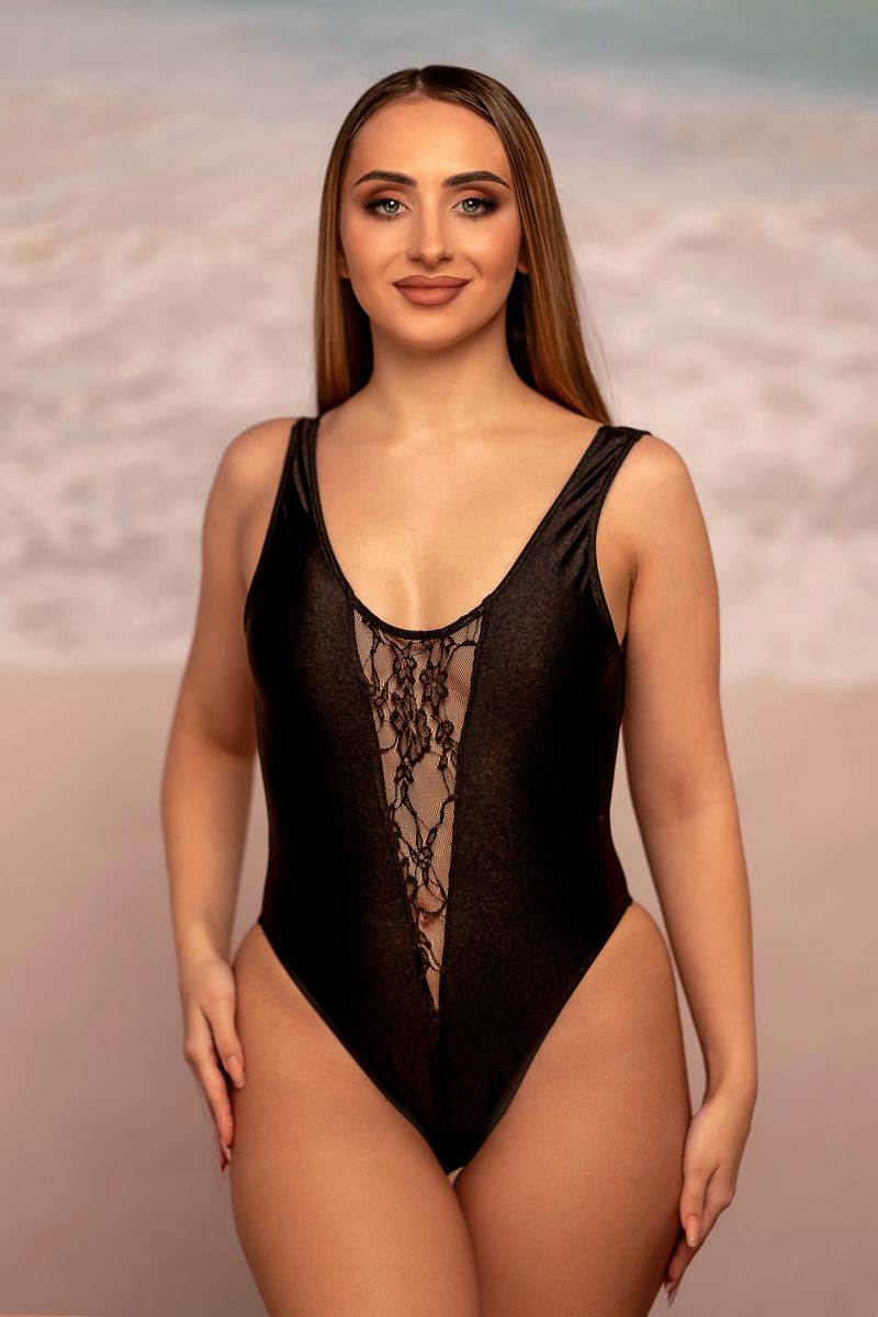 Woman wearing the Bliss One Piece by Brigitewear International, LLC, stands on a sandy beach with the ocean in the background.