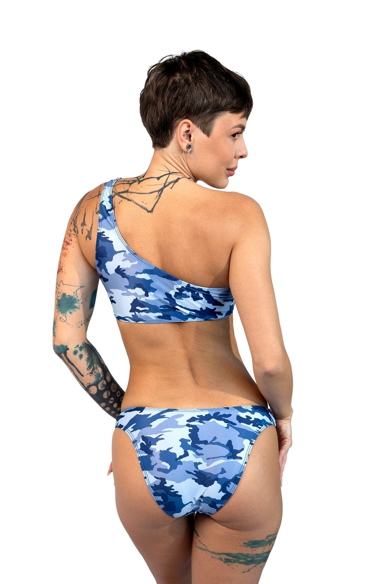Tattooed woman wearing Brigitewear International’s Cannes Thong & Rio Bikini Bottom, one-shoulder blue camouflage style, back to camera.
