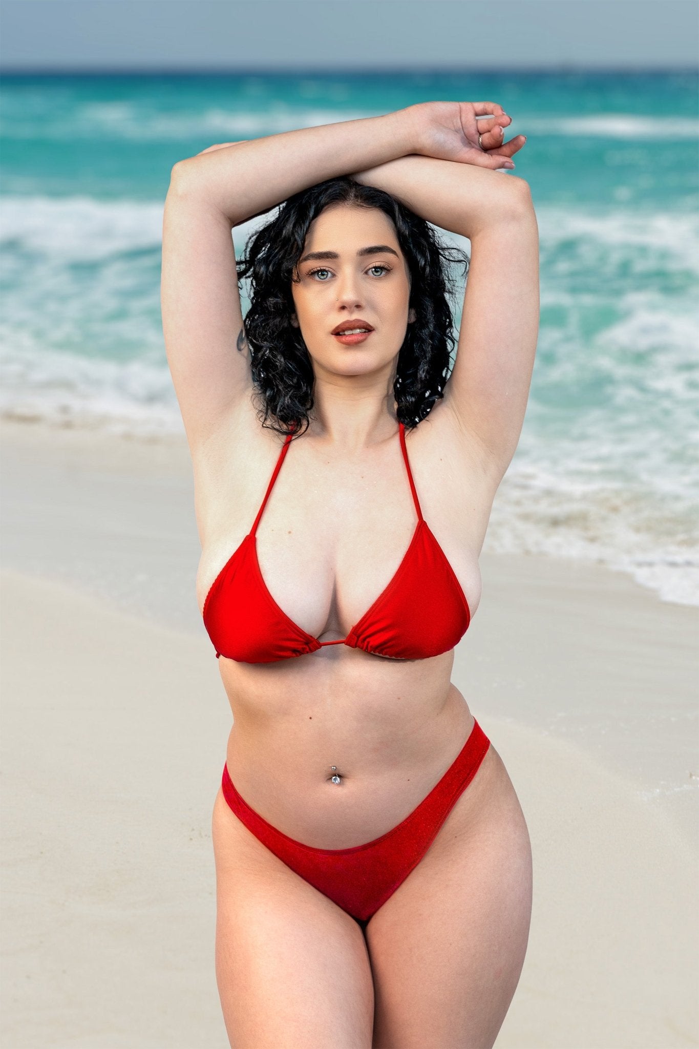 Woman wearing the Cannes Bikini Top by Brigitewear International, LLC stands on a sandy beach with turquoise ocean waves in the background.