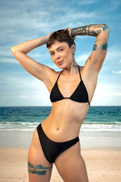A woman with short hair and tattoos poses on a beach in the Brigitewear International, LLC Cannes Bikini Top, with the ocean and sky in the background.