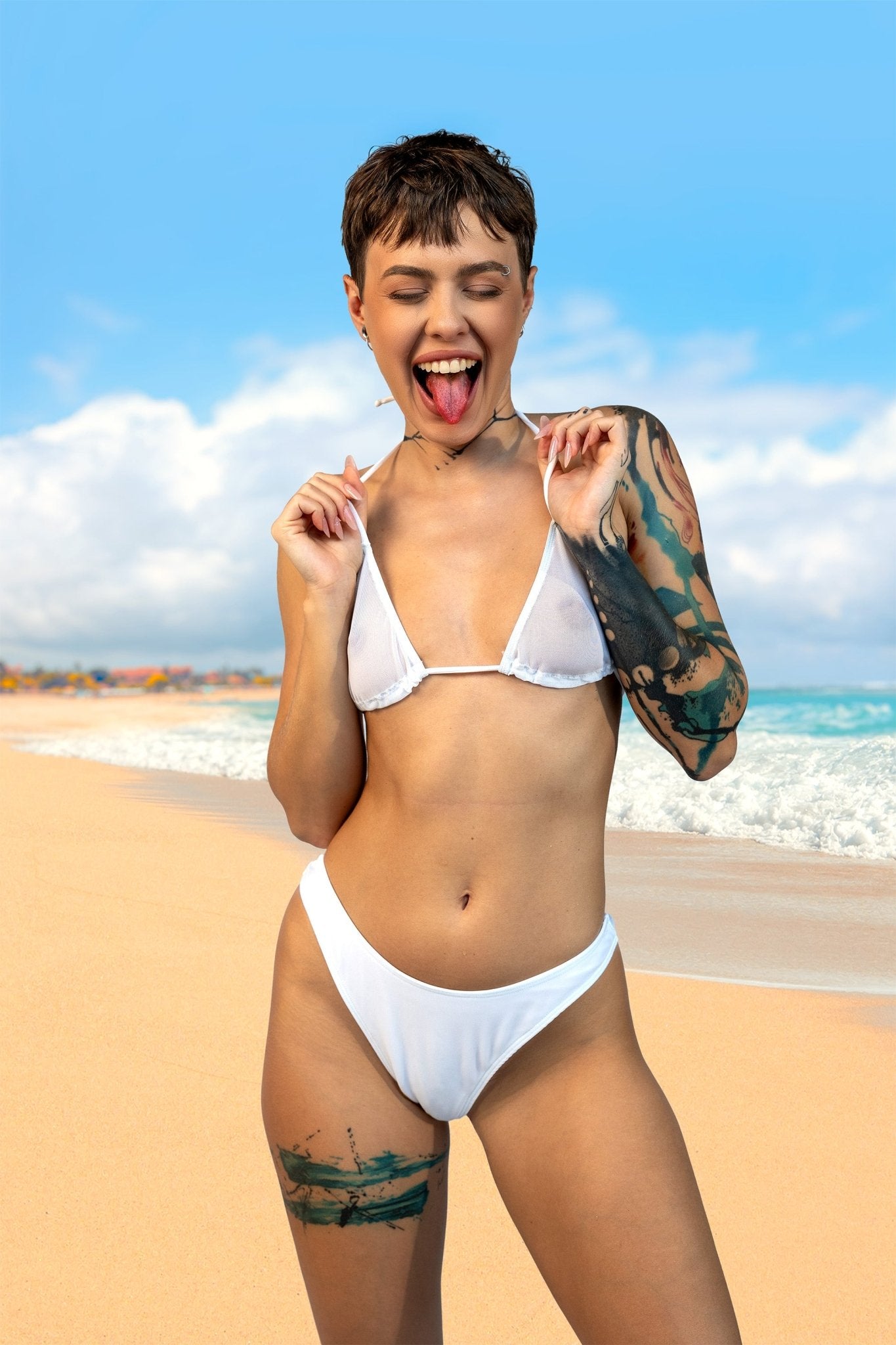 A woman smiles and poses playfully on a sunny beach with waves in the background, wearing the Cannes Mesh See Through Bikini Top by Brigitewear International, LLC.