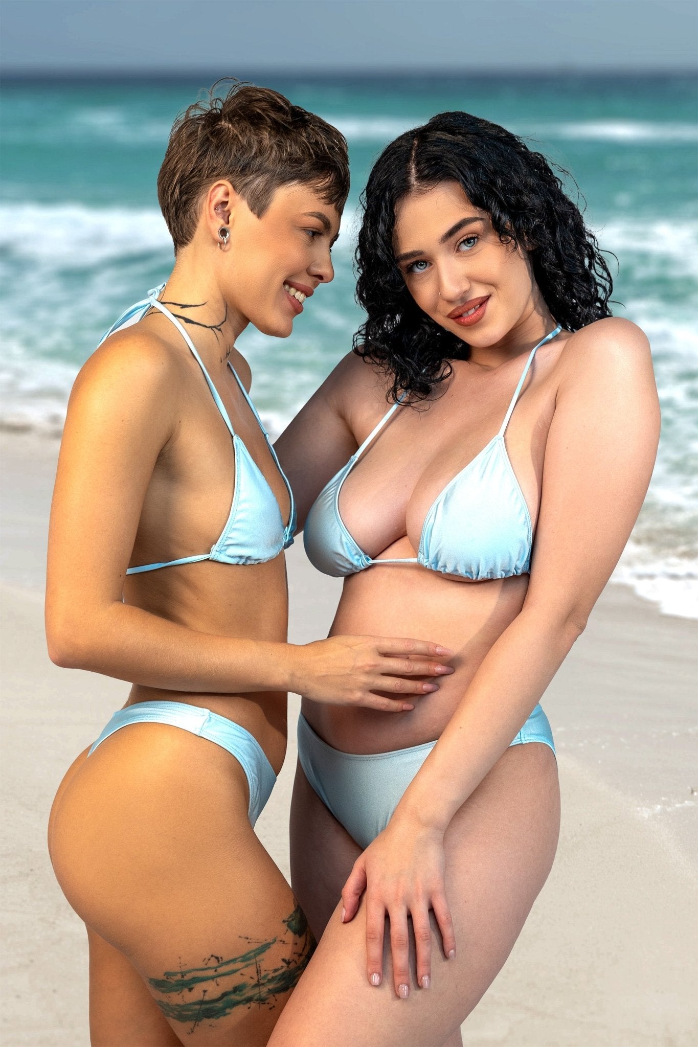 Two women in Cannes Thong & Rio Bikini Bottoms by Brigitewear International, LLC embrace and smile on a sandy beach with waves in the background.