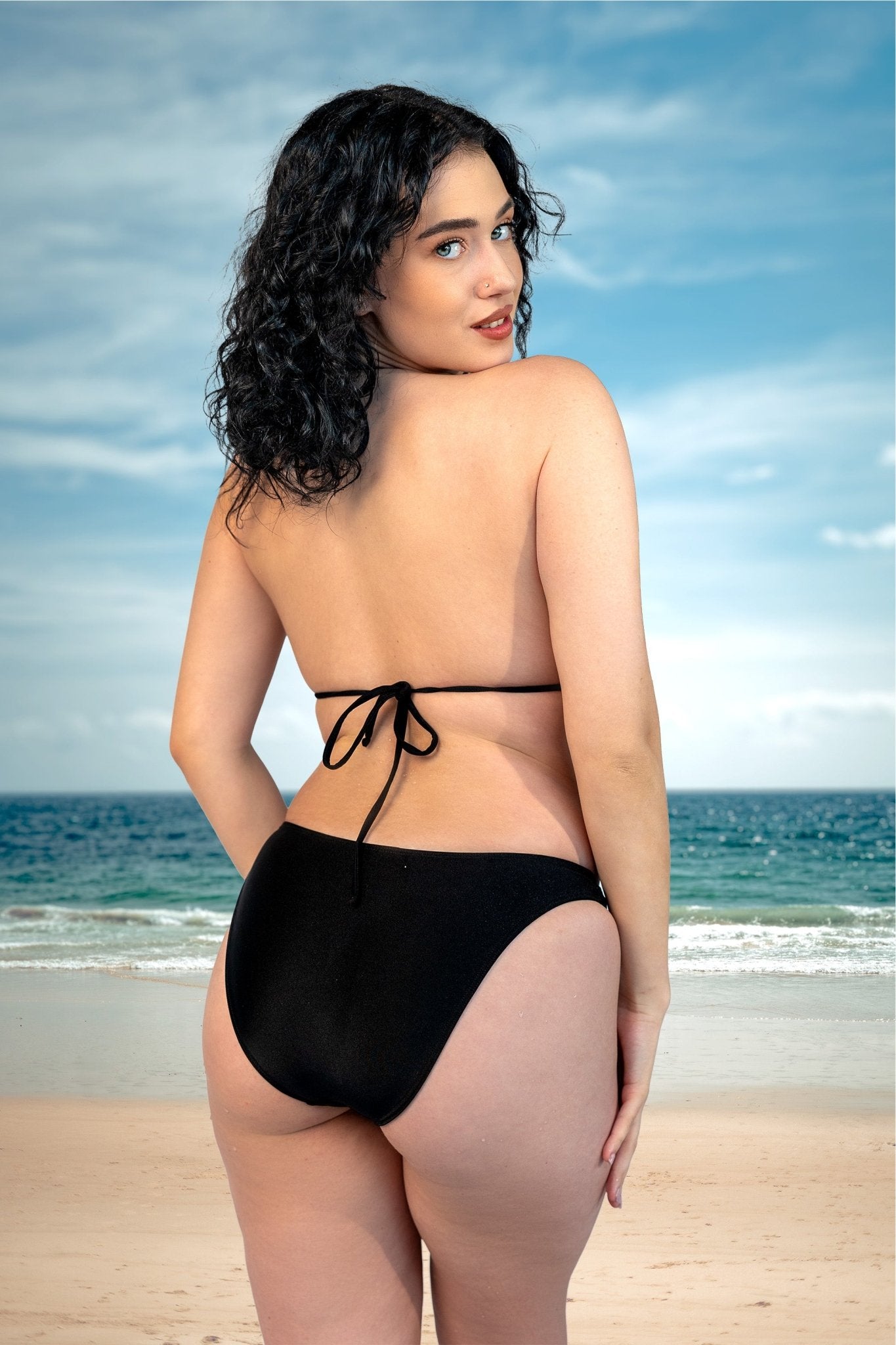 Woman with curly dark hair, wearing the Cannes Thong & Rio Bikini Bottom by Brigitewear International, LLC, stands on a sandy beach in a black bikini, facing away from the camera.