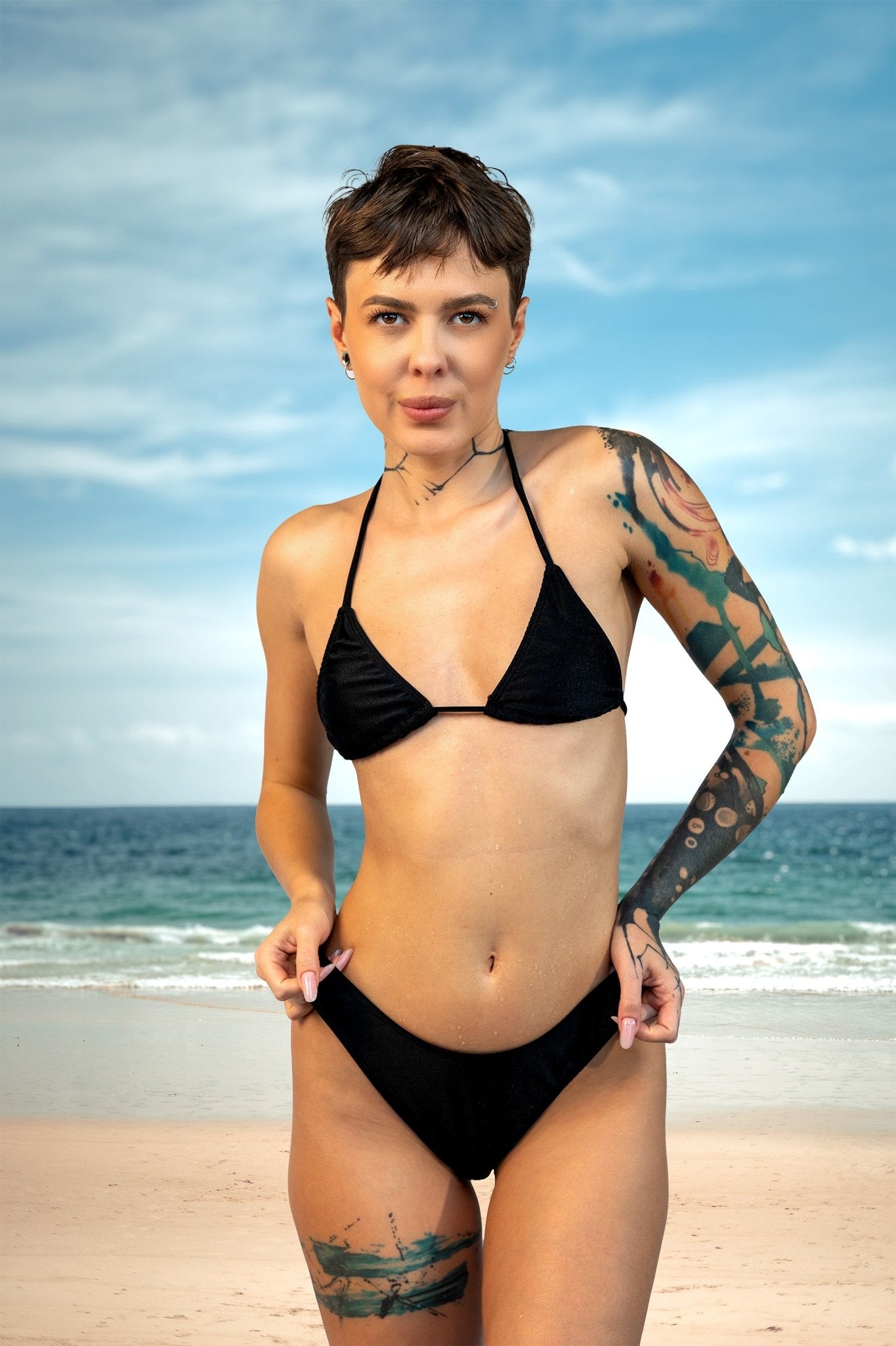 A person with tattoos wears the Cannes Thong & Rio Bikini Bottom by Brigitewear International, LLC, standing on a sandy beach with the ocean and blue sky in the background.