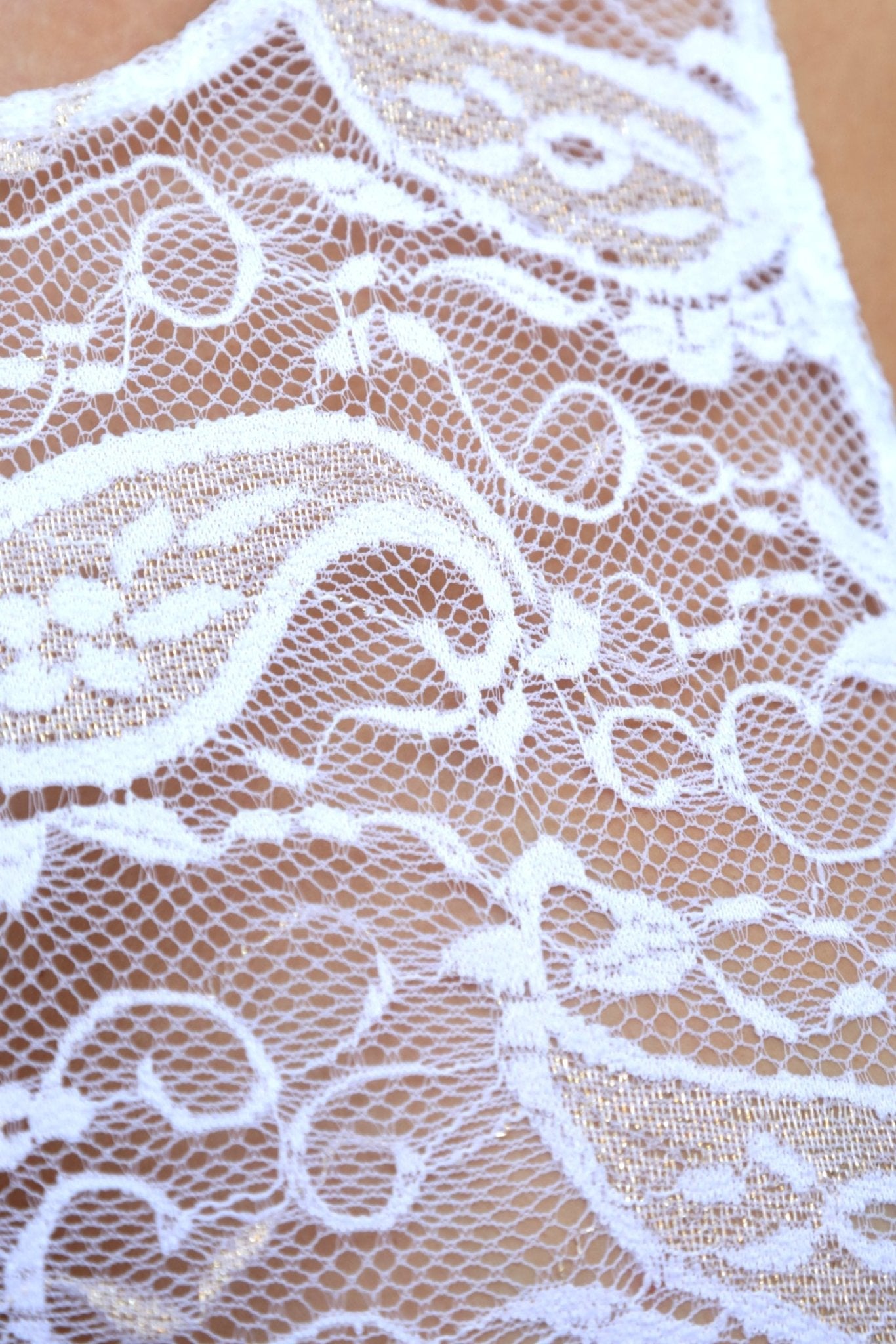 Close-up of white lace fabric with intricate floral and paisley patterns over a beige background, perfect for resort wear or as an elegant swimsuit cover-up.