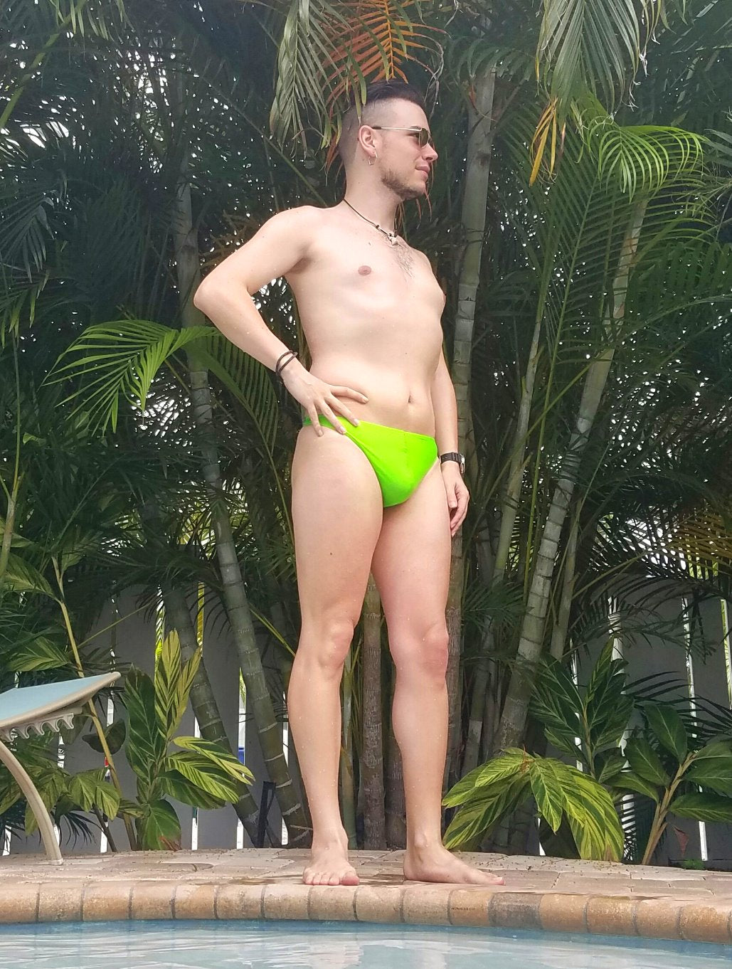 A person in a bright green swimsuit stands by a pool, surrounded by palm trees and greenery.