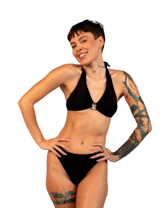 A woman with tattoos models Brigitewear International, LLC's Ecstasy Bottoms—sexy and stylish bikini bottoms—in black, posing with hands on hips against a light gray background.