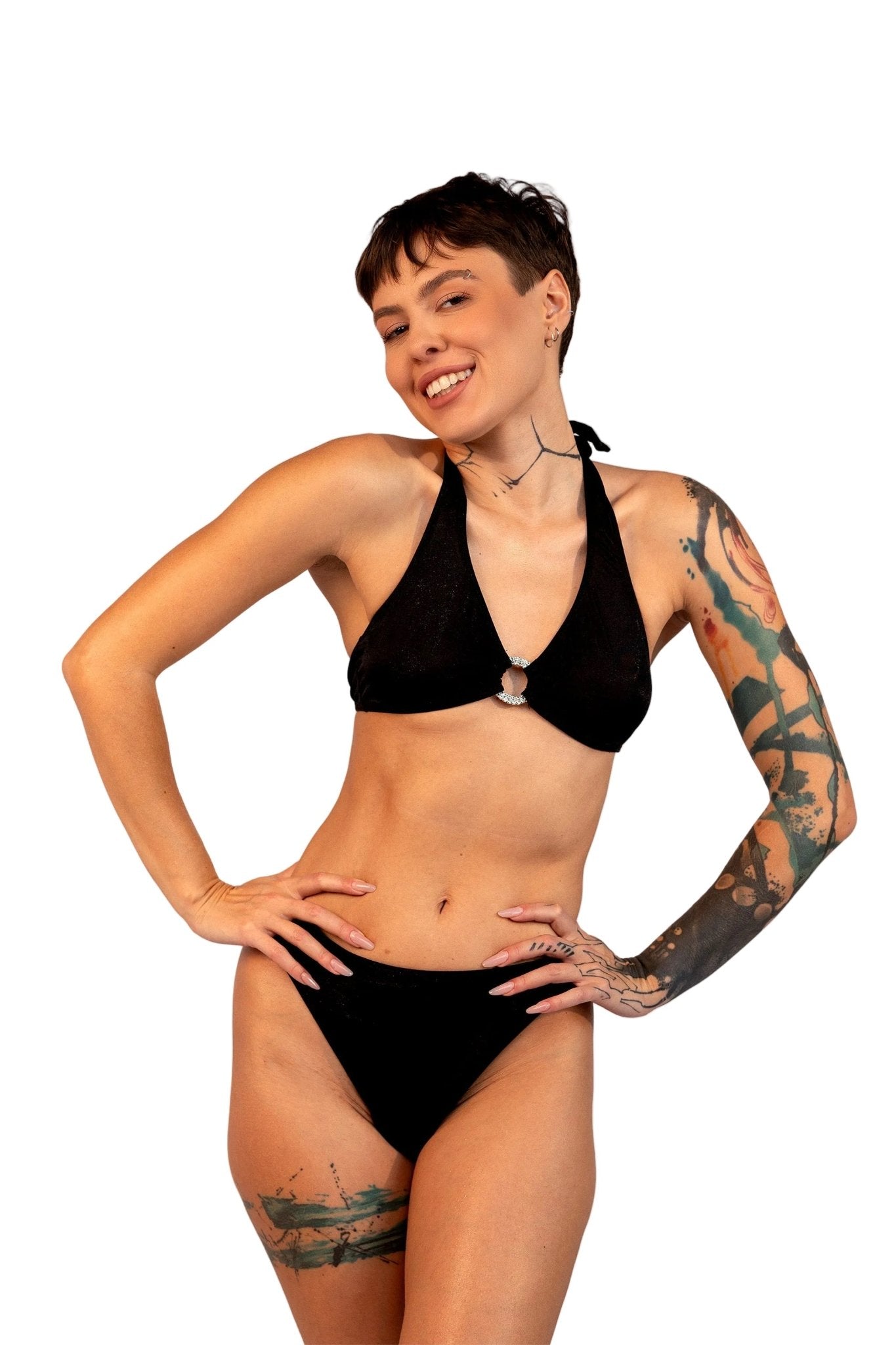 A woman with tattoos models Brigitewear International, LLC's Ecstasy Bottoms—sexy and stylish bikini bottoms—in black, posing with hands on hips against a light gray background.