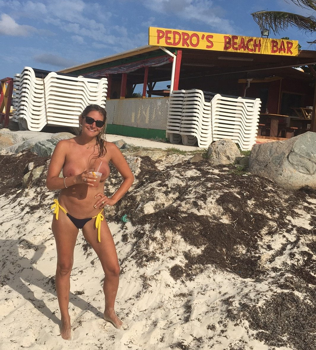 Woman in bikini holding a drink on a sandy beach in front of Pedros Beach Bar, with stacked chairs behind her.