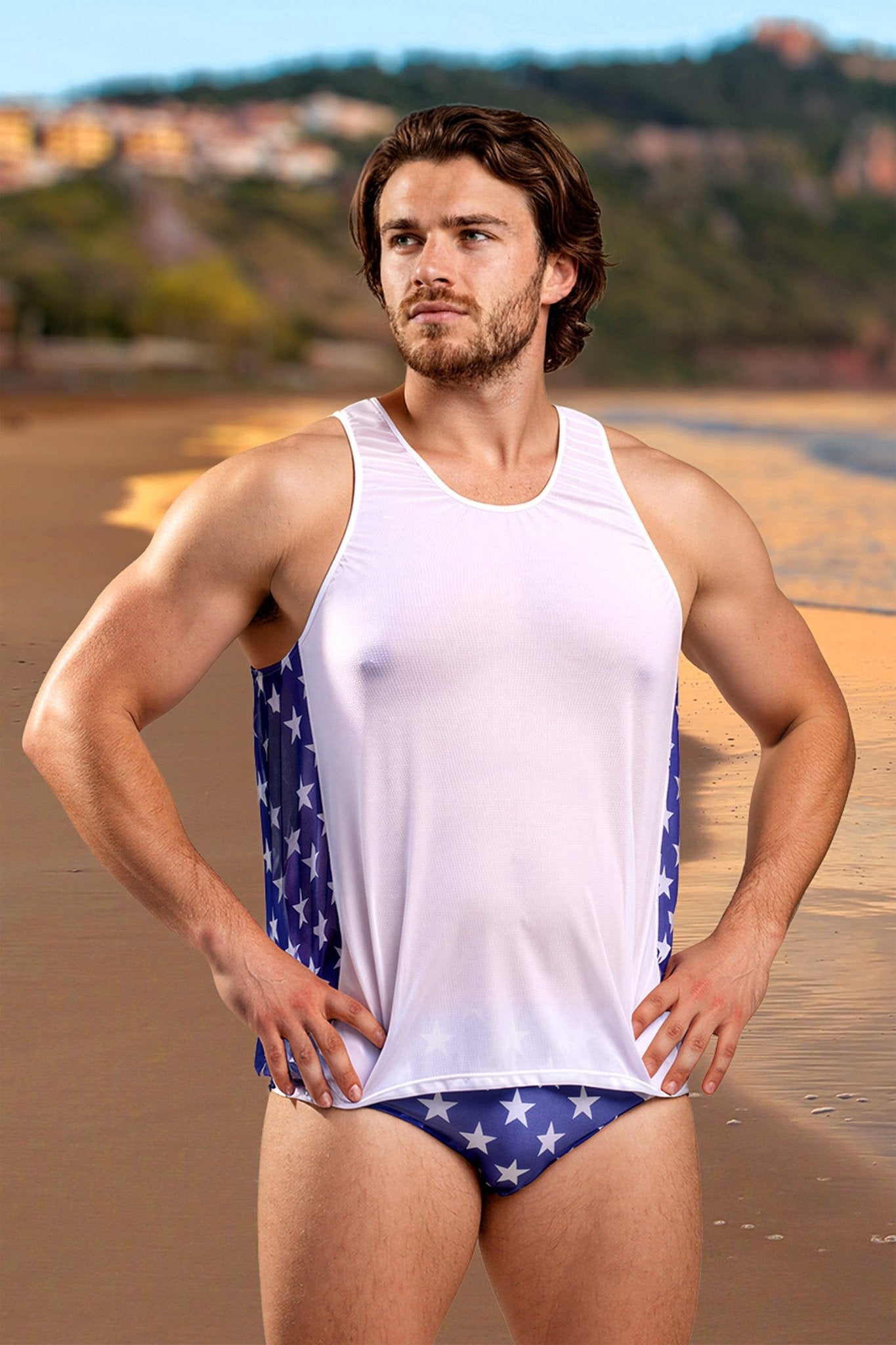 A man wearing the Independence Men's Tank Top by Brigitewear International, LLC stands on a sandy beach with buildings in the background.