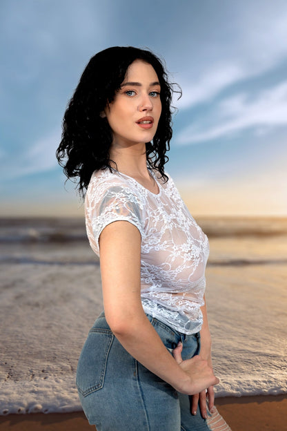 Woman with dark curly hair wears the Brigitewear International Long Sheer Lace Tee with jeans, standing on the beach at sunset and looking at the camera.