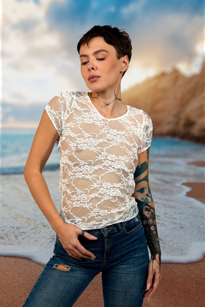 Person with short hair wears the Brigitewear Long Sheer Lace Tee by Brigitewear International, LLC with jeans, standing on a beach with ocean and cliffs in the background.