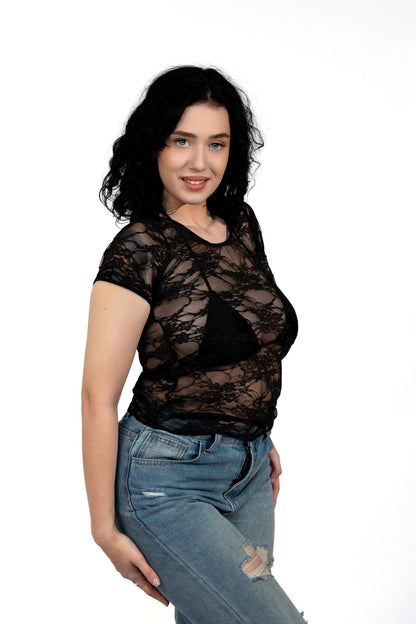 A woman with dark hair poses against a white background wearing the Long Sheer Lace Tee by Brigitewear International, LLC paired with ripped jeans.