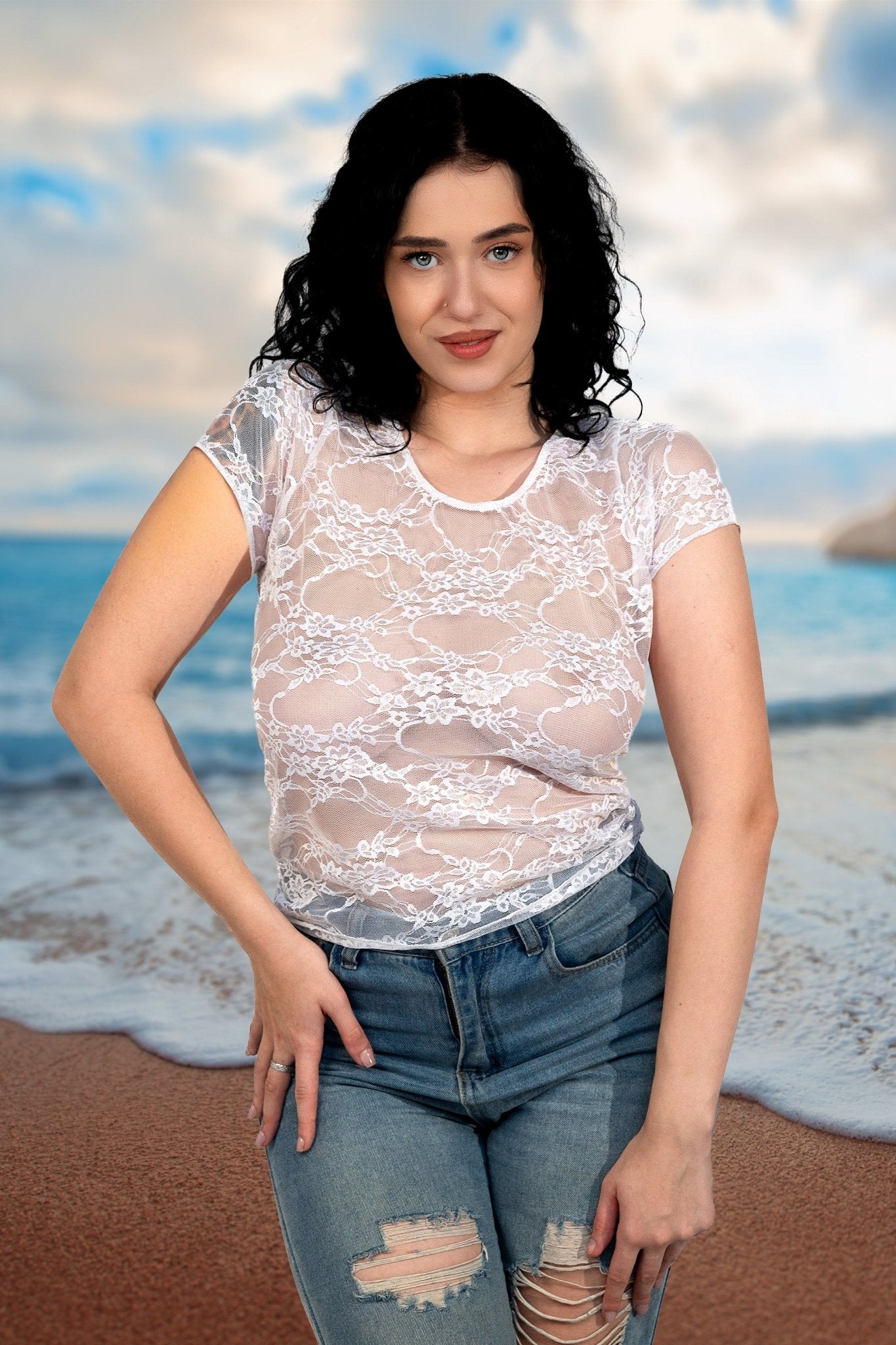 A woman with dark curly hair wears the Brigitewear Long Sheer Lace Tee by Brigitewear International, LLC and ripped jeans, standing on a beach with waves in the background.