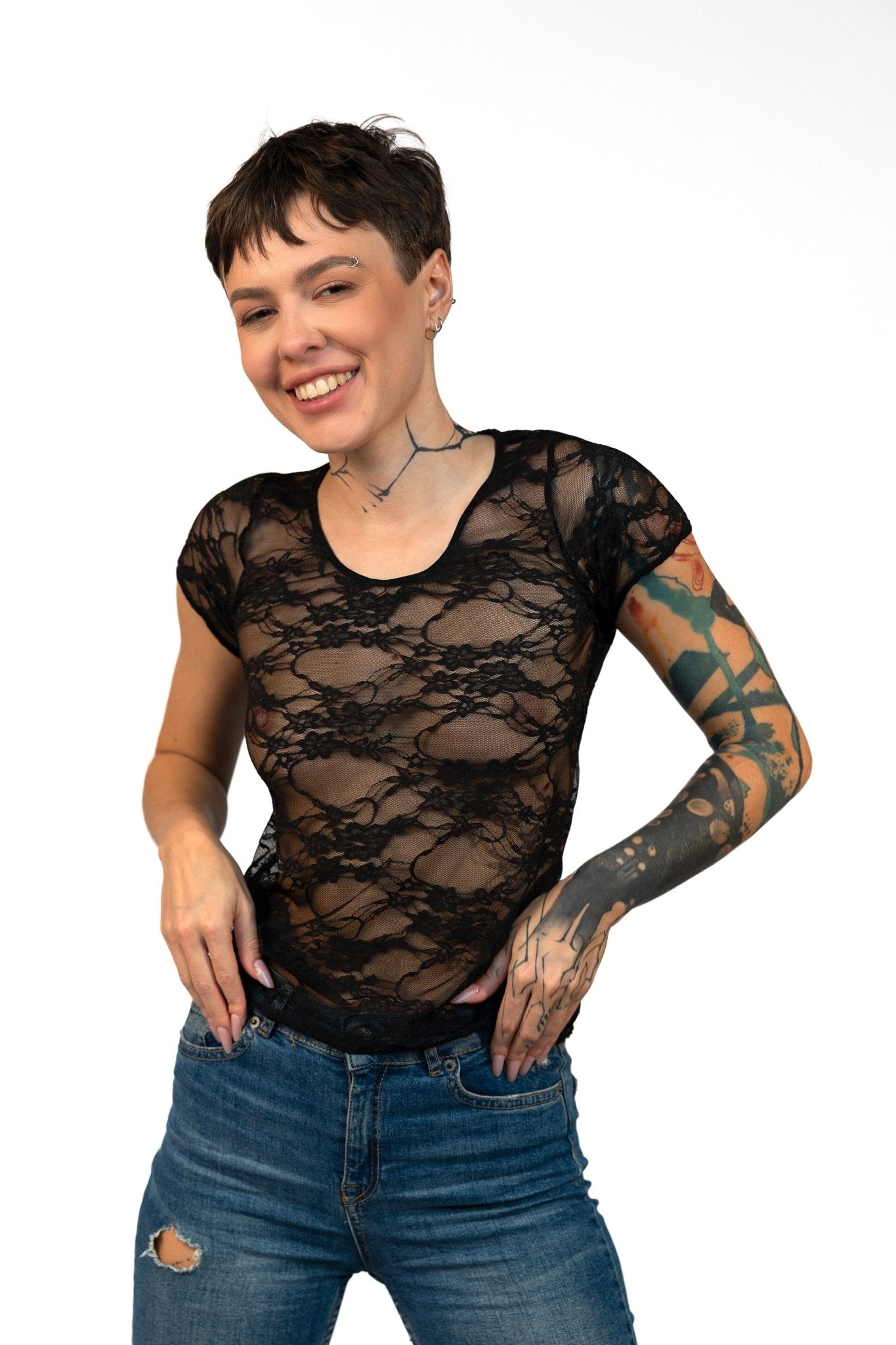 Smiling person with short hair and tattoos poses in the Long Sheer Lace Tee by Brigitewear International, LLC, hands on hips against a white background.