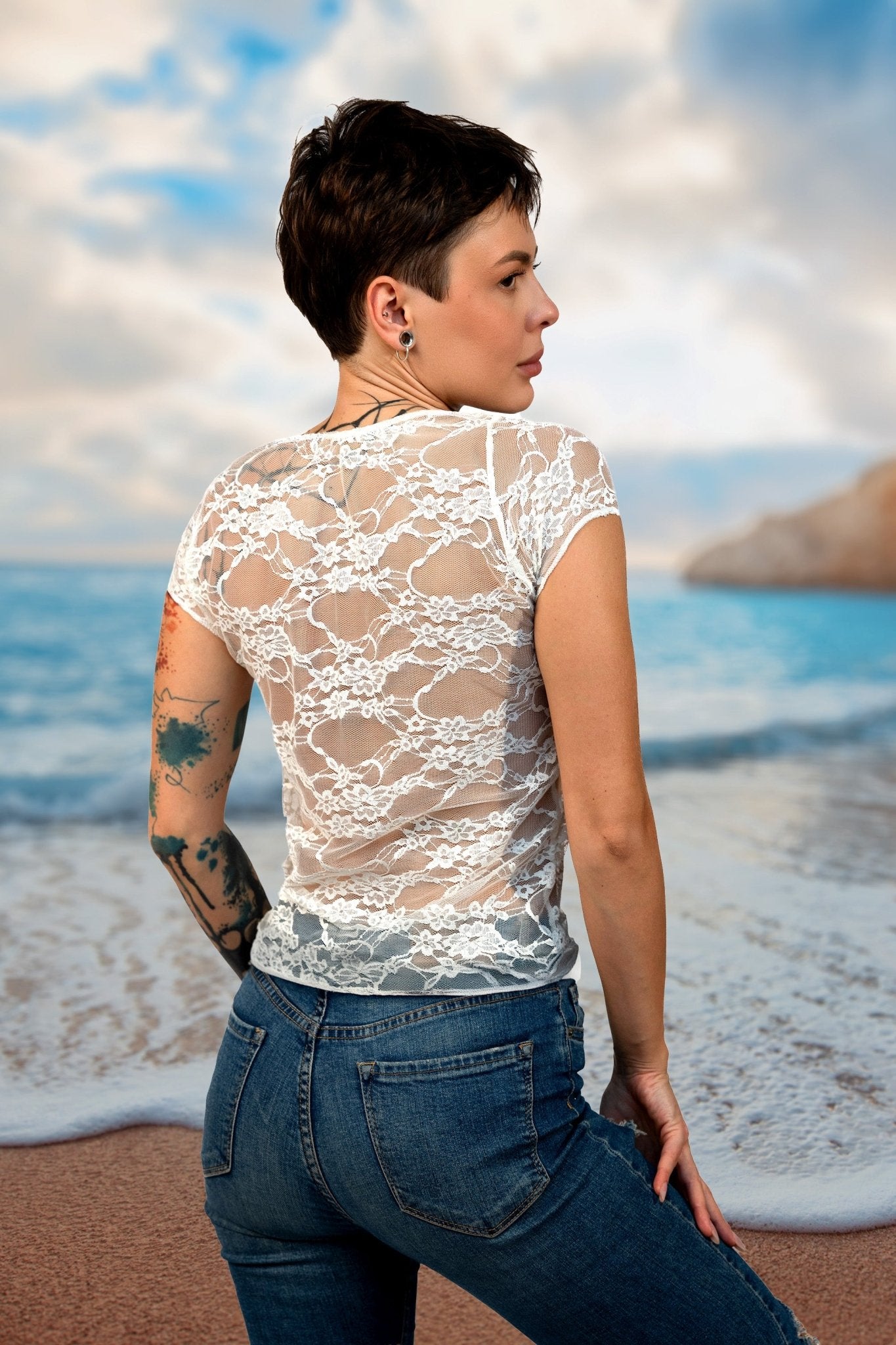 A woman with short hair and tattoos stands on a beach, facing the ocean, wearing Brigitewear International’s Long Sheer Lace Tee.