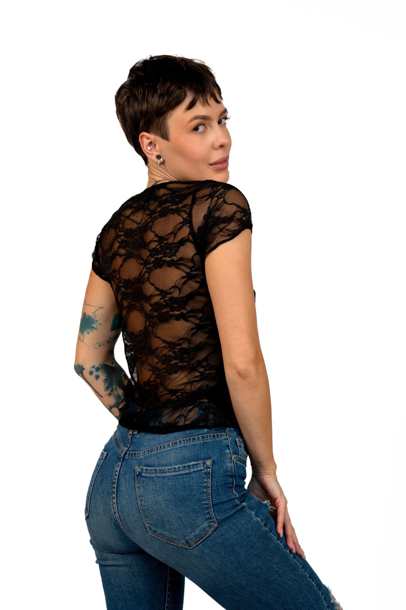 Model with short hair shows off the Long Sheer Lace Tee by Brigitewear International, LLC, pairing it with jeans and posing with her back turned while glancing over her shoulder against a white background.