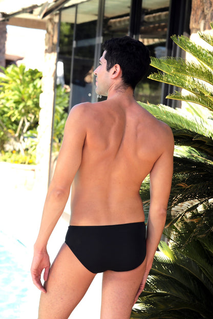 A man showcases Brigitewear International, LLC's "Men's Sheer Bravado: Bold & Confident Style" swim trunks as he stands poolside, facing away, with lush greenery and glass windows behind him.