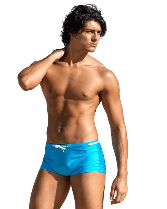Man wearing bright blue Men’s Square Cut Swim Shorts – Low Rise, Sheer & Sexy Fit standing on a rocky beach.